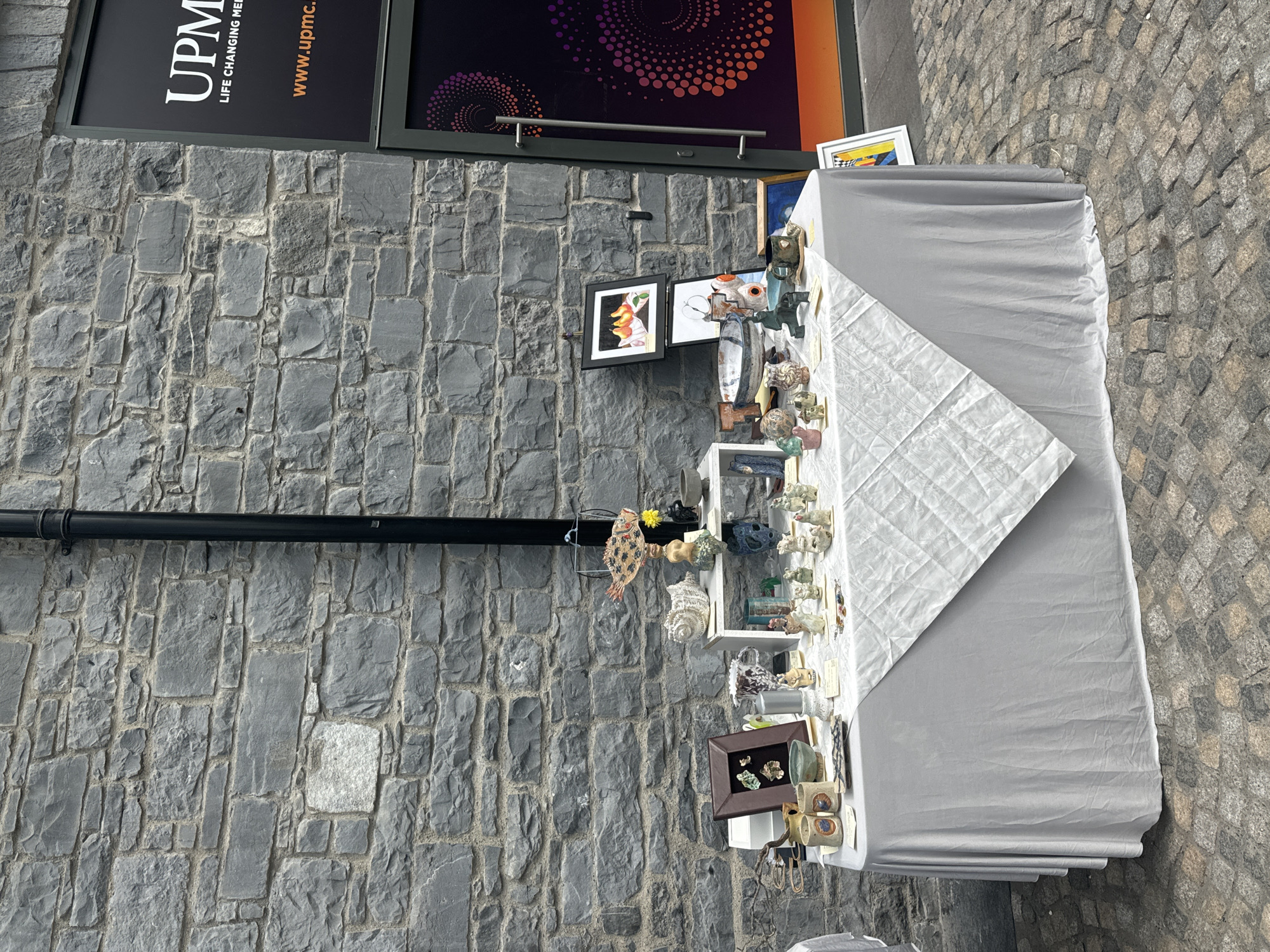 The image shows a table displaying various ceramic art pieces and framed artwork against a textured stone wall. The table is covered with a light gray tablecloth, and a white patterned runner is draped diagonally across it. On the table, there are numerous small ceramic figurines of animals, people, and abstract shapes, as well as some larger ceramic bowls and vessels. To the left of the table, there are two framed pieces of artwork, one depicting pears and another appearing to be a sketch. A sign with the UPMC logo and text "LIFE CHANGING MEDICINE" and "www.upmc.ie" is visible on a dark-colored building to the right of the scene. The ground is paved with cobblestones. The overall setting suggests an outdoor market or exhibition. This scene is located in front of the UPMC building on the Kilkenny city Medieval Mile, Ireland.