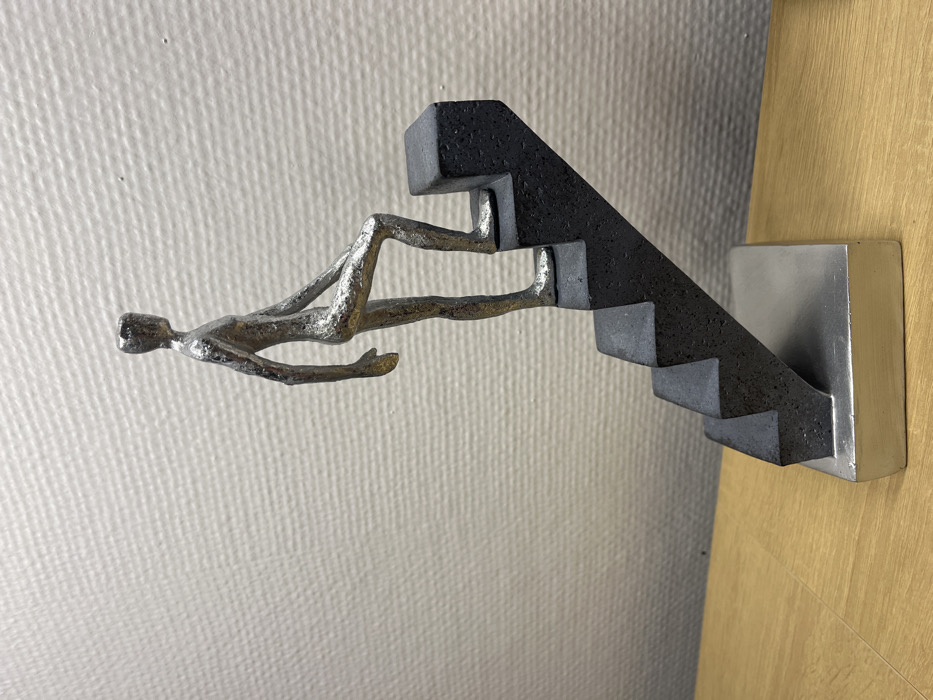 An indoor scene displays a metallic sculpture situated on a light brown wooden surface, positioned against a light gray wall with a subtle textured pattern. The sculpture features a stylized, silver-colored human figure, rough in texture and lacking distinct facial features, depicted in the act of ascending a dark gray, five-step staircase. The figure's right foot rests on the second step from the top, with its left foot on the step below, conveying a sense of upward movement. The dark gray staircase structure is secured to a rectangular silver-colored base. The wooden surface exhibits visible grain, and the background wall presents an even, fine texture. While the location is indicated as Schönau am Königssee, Germany, the image contains no discernible visual elements to confirm this specific geographic placement.