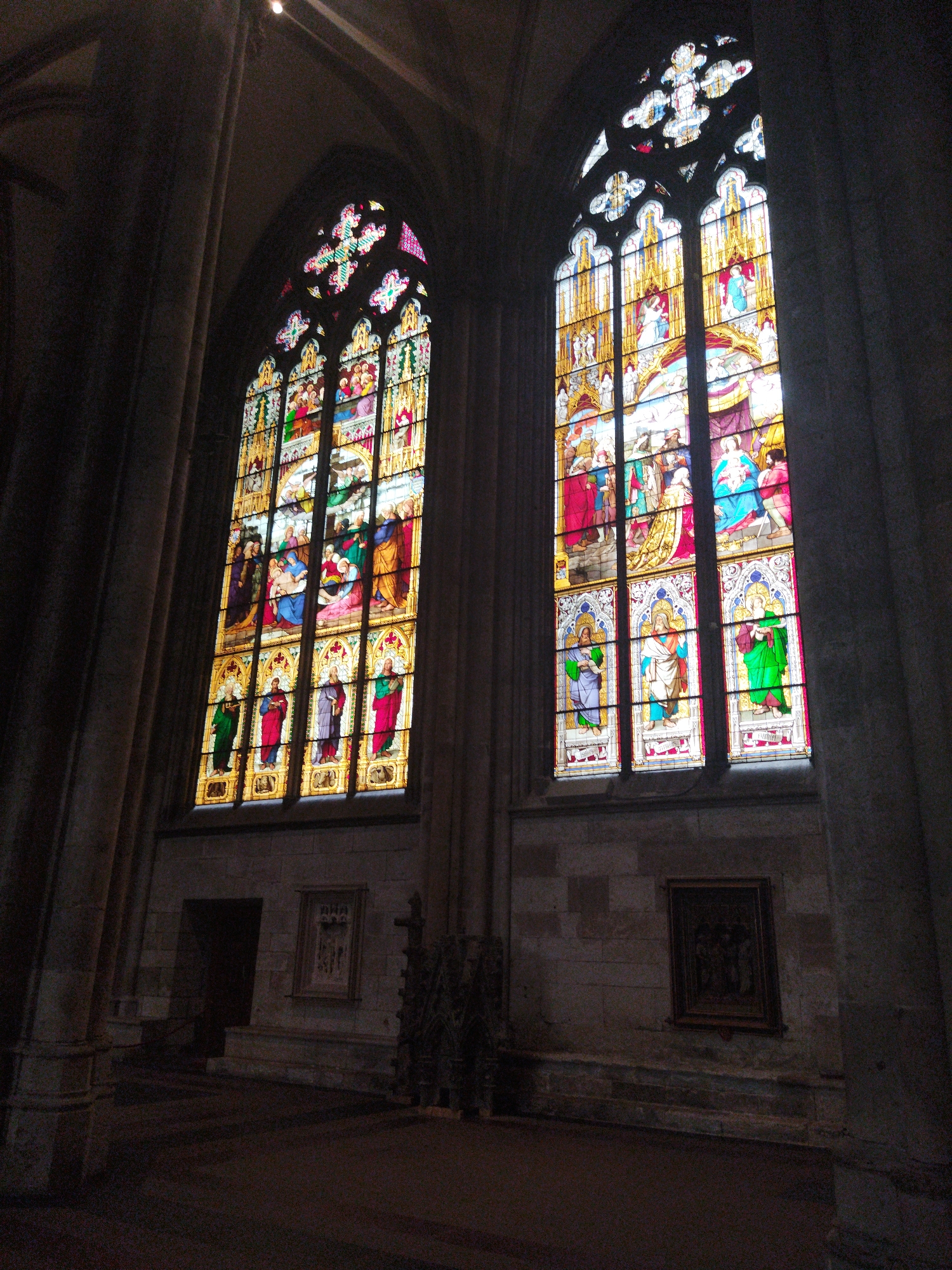 The image shows a view of the interior of St. Gereon's Basilica in Monheim am Rhein, Germany. The focus is on two large, ornate stained-glass windows in a Gothic architectural style. The windows are tall and arched, divided into multiple panes, each depicting colorful scenes with religious figures. The left window is partially obscured by shadow, but the right window is more fully illuminated, showing detailed figures in robes and elaborate headdresses.
The windows are set within a stone wall of the basilica, with a large, dark pillar on the right side of the frame. In the foreground, the floor is dark and appears to be stone. To the left of the windows, there is a dark archway and a smaller framed artwork on the wall. To the right of the windows, another framed artwork hangs on the stone wall. The overall lighting is dim, with light filtering through the stained glass, creating a somber and awe-inspiring atmosphere typical of a historic church interior.