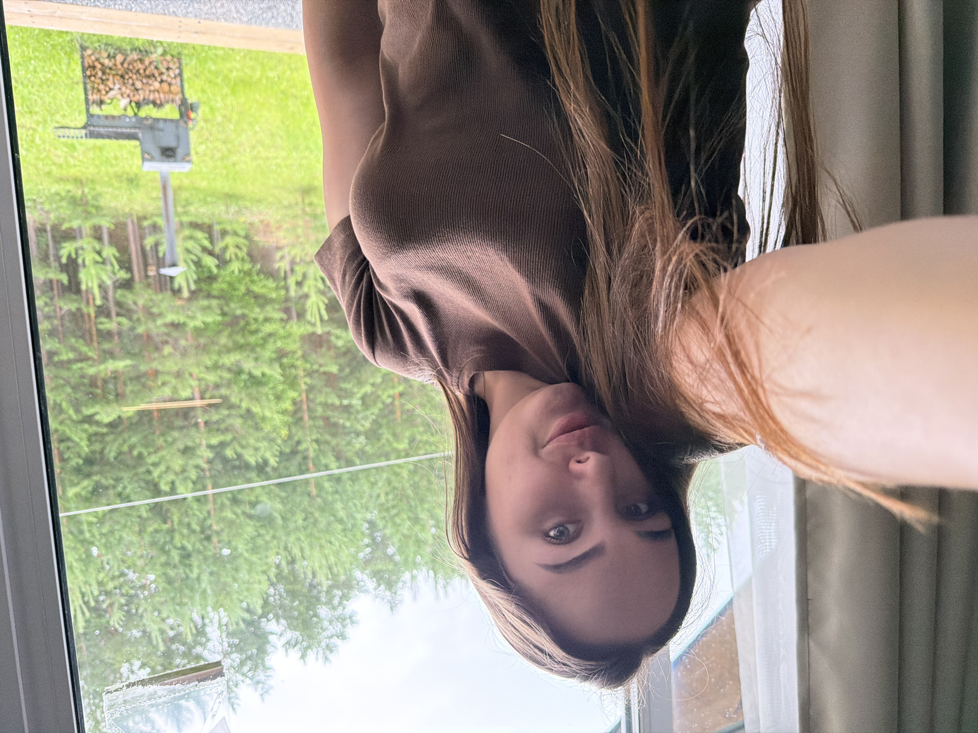 A young woman with long, brown hair is taking a selfie in front of a window overlooking a wooded area. She is wearing a brown, ribbed t-shirt. Through the window, pine trees and a green, grassy field are visible. There is also a metal grill with a stack of firewood next to it. The scene appears to be in a rural or suburban location, possibly Kamyennoye, Russia, given the presence of pine trees and a rustic outdoor setting. The sky is overcast.