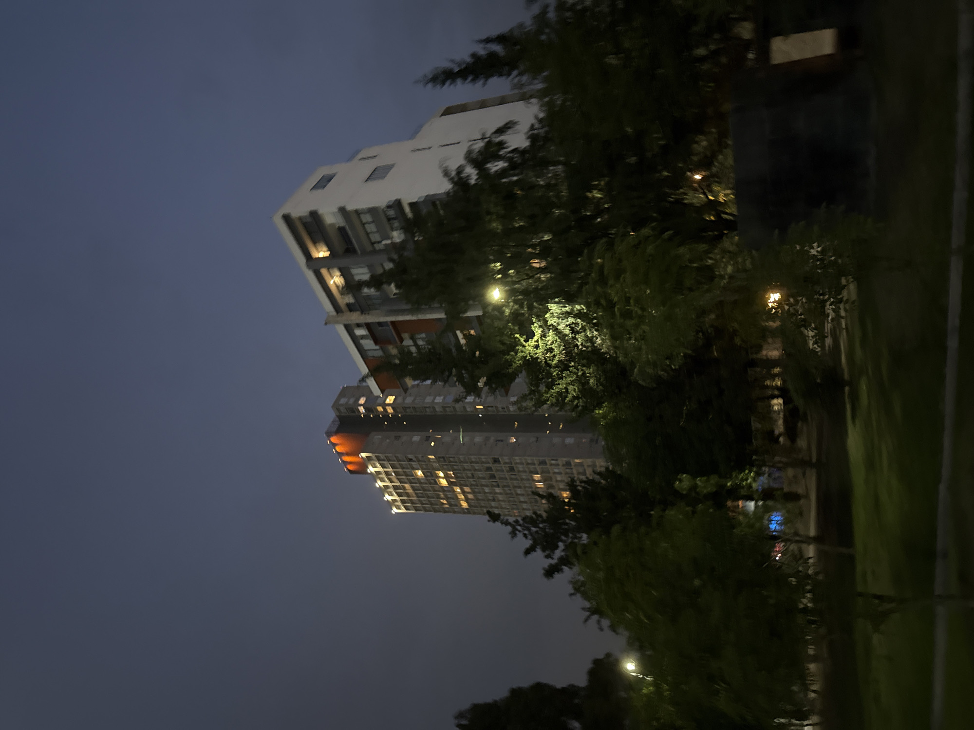 The image shows two buildings, one white and the other gray, lit up at night in Puebla, Mexico. A dark, overcast sky fills the background. Trees and greenery surround the buildings, obscuring parts of their facades. Streetlights illuminate the foreground.