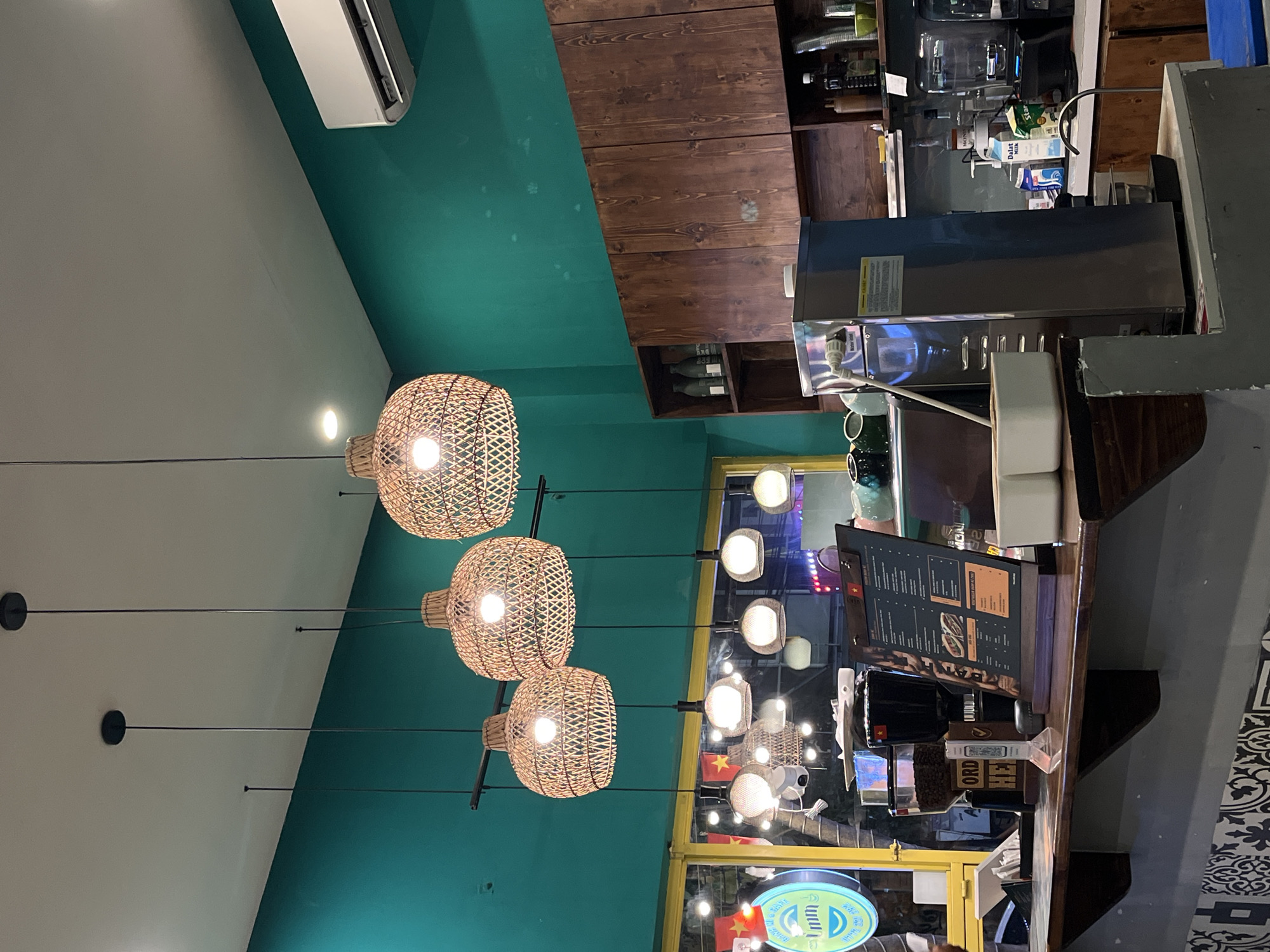 The image depicts the interior of a cafe or restaurant, possibly in Đà Nẵng, Vietnam. The focal point includes three hanging pendant lights with woven shades, casting a warm glow. Overhead, there is a dark wood cabinet or shelf, and an air conditioning unit is visible on the upper left. A counter area is on the right side of the image, where a menu board is placed. Visible through a window are more lights and what appears to be a Vietnamese flag.