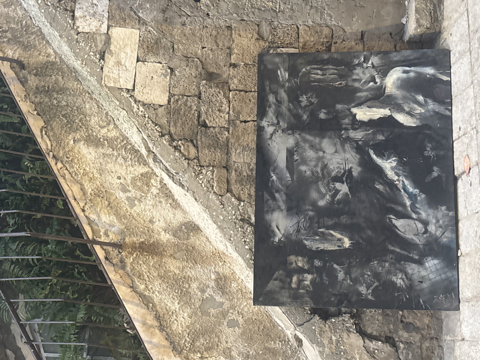 The image captures an overhead view of a dark, abstract painting resting on a rugged outdoor surface. The photograph is oriented such that the painting occupies the right half of the frame, lying flat on the ground.
The ground surface is divided diagonally. The upper left and lower left portions consist of a light-colored, textured concrete or plaster-like material, showing signs of age with cracks and uneven patches. Towards the bottom left, a dark metal railing or fence structure is partially visible, with green foliage peeking out from behind it, suggesting a drop-off or a garden area adjacent to the path. The upper right and lower right parts of the ground are composed of an irregular stone wall or paved area, made of varied sized, light-colored rectangular and square stone blocks, cemented together with visible, coarse mortar.
The painting itself is rectangular, with a dominant palette of black, dark gray, and white, with subtle hints of other colors. It features a dynamic and somewhat chaotic composition, suggestive of movement and form within darkness.
In the upper right section of the painting, a prominent, light-colored form resembling a bird, possibly a swan or dove, is depicted with outstretched wings, appearing to be in flight or soaring. This form is rendered primarily in white and light gray, contrasting sharply with the darker background.
Below and to the left of this form, the painting dissolves into swirling, textured brushstrokes. There are areas of smeared white and light gray paint against the dark canvas, creating a sense of depth and motion. Other indistinct forms and shapes emerge from the darker areas, contributing to an abstract, almost dreamlike quality. A small, dark vertical rectangle is visible in the upper-middle left portion of the artwork. Towards the bottom right corner of the painting, a small grid-like pattern can be discerned.
In the very bottom right corner of the painting, a signature or inscription is visible. While somewhat stylized and partially obscured by the texture of the painting, it appears to read "F.K.J" or "F.K.I" followed by additional indistinct characters or a symbol.
The lighting suggests natural daylight, possibly indirect, as shadows are soft and textures on both the ground and the painting are clearly delineated. The overall impression is one of an artwork being displayed or photographed in an old, possibly historic outdoor setting. No people are visible, and the scene is static.