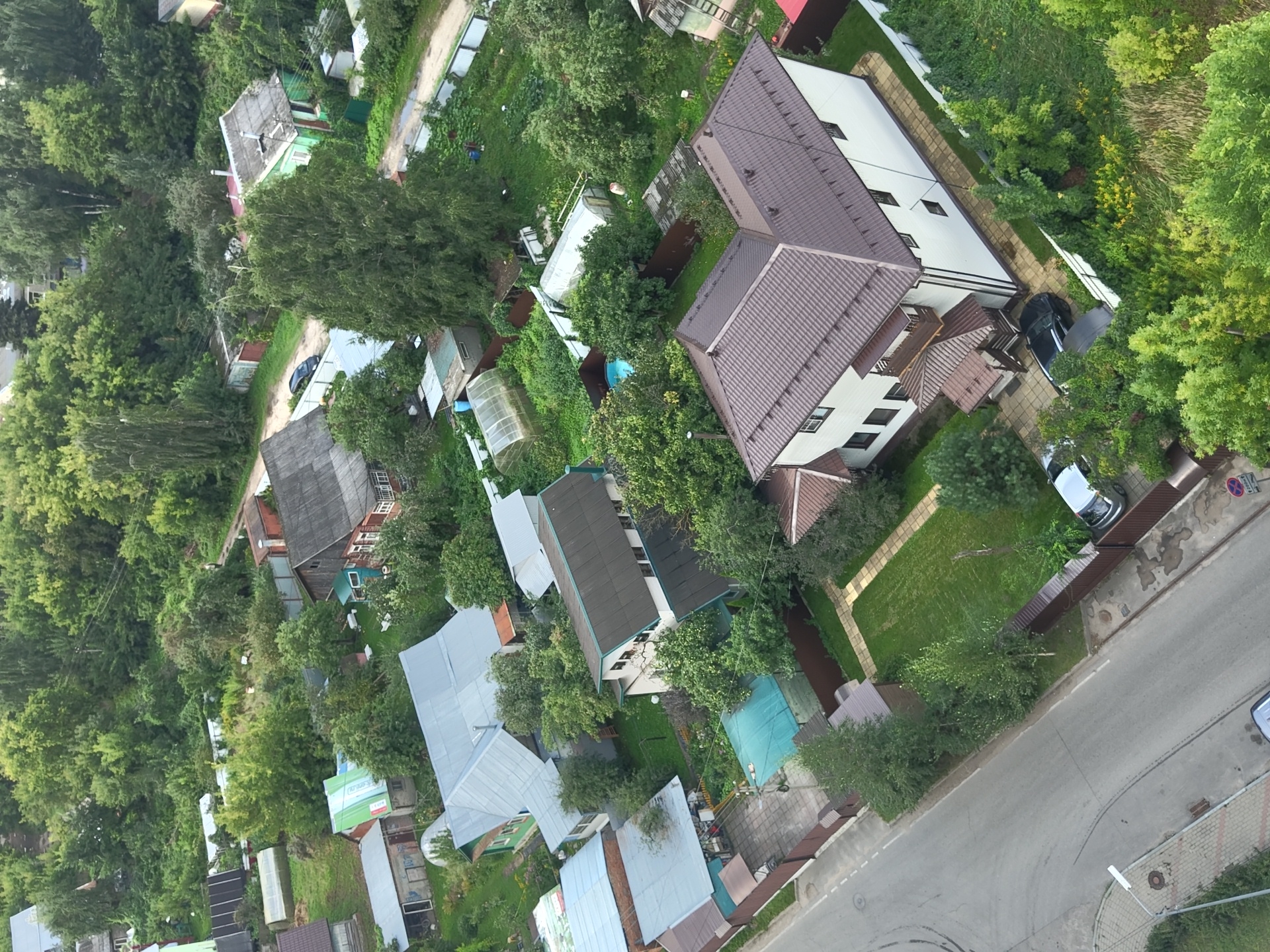 This is an aerial view of a suburban residential area in Ramenskoye, Russia, likely taken on a cloudy day. The scene is dominated by numerous houses with pitched roofs, many featuring metal roofing in various colors, including gray and brown. The properties are interspersed with abundant trees and lush greenery, indicating a well-established neighborhood with mature landscaping.
Several houses are visible, ranging from single-story structures with garages to larger, multi-story dwellings. One prominent house in the foreground is a two-story white building with dark trim and a dark brown, multi-pitched roof. It has several windows and a visible balcony. Other houses are less distinct due to vegetation cover and distance.
The area is accessed by paved roads and dirt paths, with some gravel driveways leading to the houses. A few vehicles are present: a dark-colored car parked in front of the large white house, and another silver car on the road near the fence line. One property features a small blue swimming pool partially covered by a tarp. Several greenhouses are also scattered throughout the properties, suggesting gardening is a common activity.
Fencing is prevalent, with wooden and metal fences demarcating property lines. The presence of streetlights and what appear to be paved sidewalks along the main road indicates urban infrastructure. The overall impression is one of a tranquil, densely vegetated residential district.