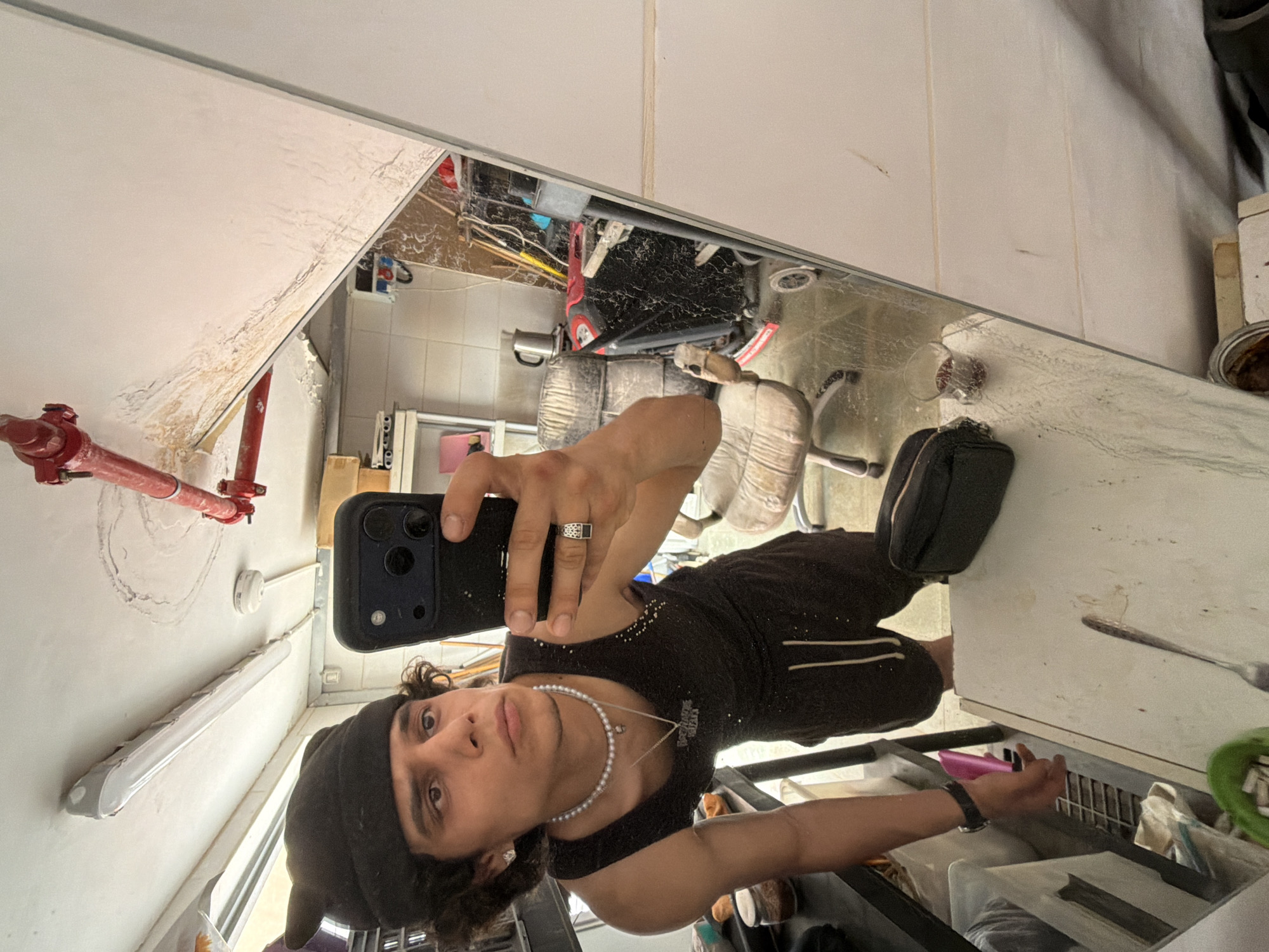 A young man takes a selfie in front of a large mirror, holding a black smartphone with his right hand. He wears a black beanie, a dark tank top, a white pearl necklace, and a silver ring with a dark stone on his left ring finger. A black wristwatch is visible on his left wrist. He has a neutral expression, looking directly into the camera.
The mirror reflects a cluttered indoor space. Above the man's head in the reflection, a white ceiling or wall shows signs of discoloration and texture, possibly water damage. A red fire suppression pipe system is visible on the left side of the reflected background, running horizontally along the wall. Below it, a long, white fluorescent light fixture is mounted on the wall.
The background visible in the mirror is disorganized, containing various objects. These include two light-colored upholstered chairs, a red object (possibly a child's ride-on toy or small machinery), and general clutter like wires and boxes. On the right side, a white appliance, resembling a refrigerator or cabinet, is visible, with a fork resting on it and a pink object near its base. A glass or cup containing dark contents is also seen on a surface. The environment suggests a functional, possibly industrial or storage area, rather than a typical residential setting. The overall lighting indicates daytime. The location is within the Hof HaCarmel Regional Council, Israel.