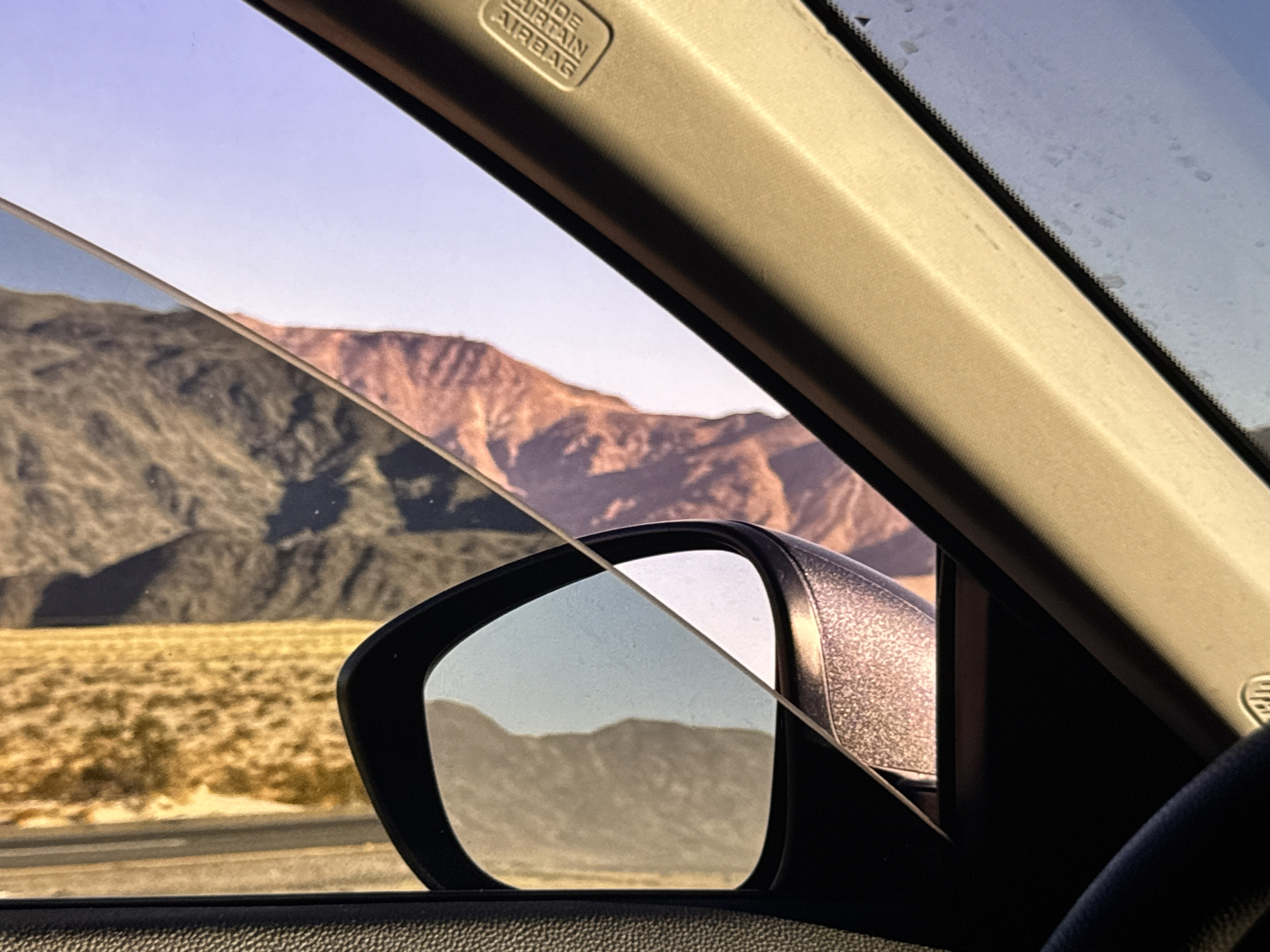 This image is a close-up view from inside a vehicle, looking out the window. The scene captured is a desert landscape with mountains in the background. A side-view mirror is visible, reflecting a portion of the same landscape. The interior of the car, including part of the dashboard and a steering wheel, is also in the foreground. The lighting suggests it is either sunrise or sunset, casting warm tones on the mountains. The image does not contain enough information to determine a specific location within Los Angeles, or any city.