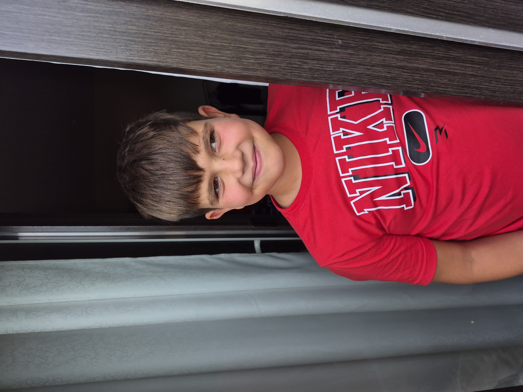 A young boy with brown hair and a red Nike t-shirt stands in a doorway, peeking from behind a dark brown wooden frame. A light-colored curtain is visible to the left of the boy, and a dark opening is behind him. He is smiling and looking to his left. The image appears to be taken indoors or in a sheltered outdoor space.