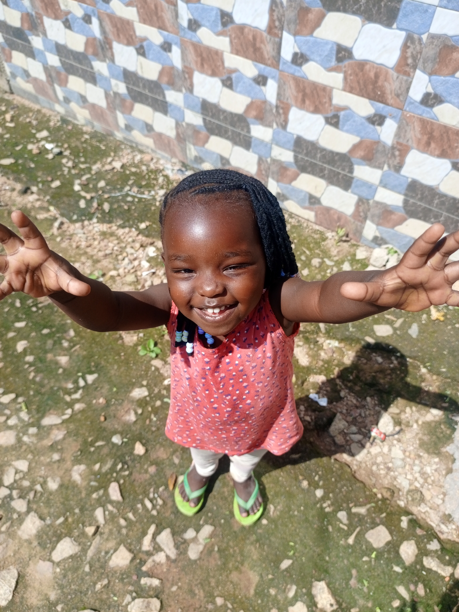 A young Black girl, likely under the age of five, with dark skin and braided hair, smiles at the camera with her arms outstretched. She wears a red and white polka dot sleeveless top, white leggings, and green flip-flops. She has a beaded necklace with white, blue, and black beads. The girl is standing on a dirt path with scattered rocks and some sparse green vegetation. Behind her is a wall tiled with an irregular pattern of brown, blue, and off-white rectangular and irregular shapes. The scene appears to be outdoors in a casual urban or semi-urban environment. The lighting suggests it is daytime, and a shadow of the girl is visible on the ground to her right. This setting is consistent with a residential area in Jos, Nigeria.