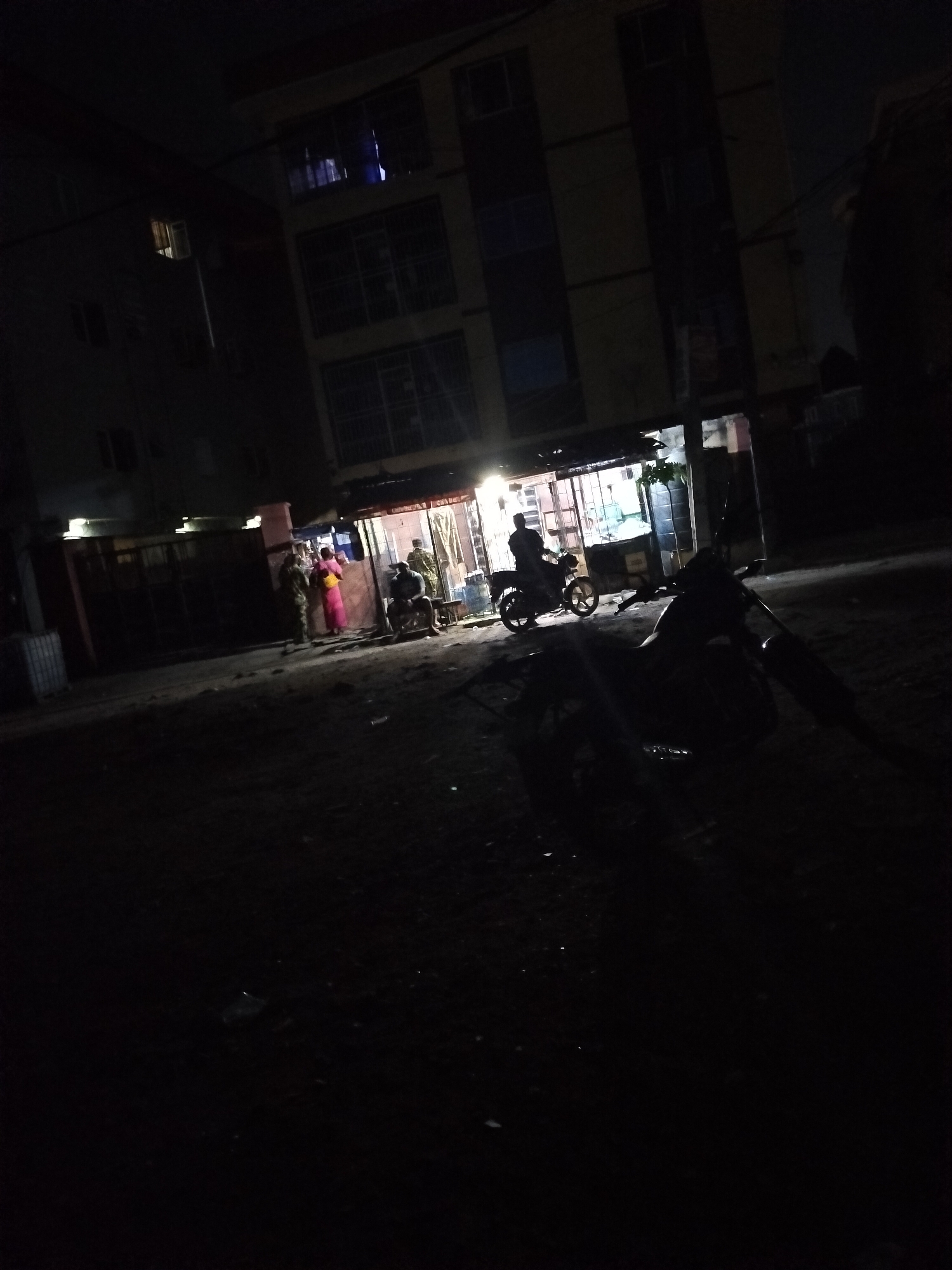 The image shows a street scene at night in Alaiyabiagba, Nigeria. The area is poorly lit. There are several buildings lining the street. A small store or shop is illuminated, with people standing and sitting nearby. Two motorcycles are visible, one parked closer to the camera in silhouette and another parked by the shop. People are engaged in conversation and activity around the shop. The road is dark and appears to be paved. There are no obvious signs of traffic or other businesses.