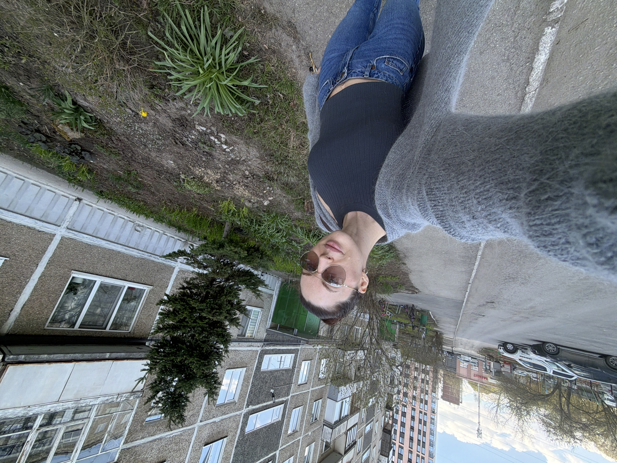 A woman wearing a black top, jeans, and a gray cardigan poses in front of a building in Perm, Russia. She wears round sunglasses. The building is multi-storied with gray concrete panels and numerous windows. A green tree grows beside it. In the background, a newer pink high-rise building is visible. A sidewalk runs along the side, and cars are parked nearby. The area appears to be a residential neighborhood.