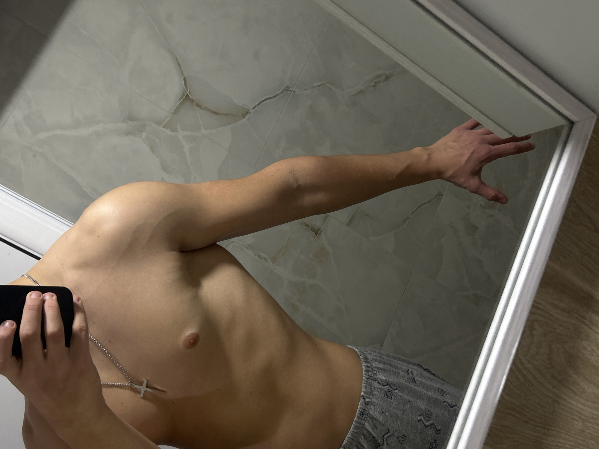 The image presents a mirror selfie of a male subject, visible from the upper chest to the upper thighs, in an indoor setting. The subject is shirtless, displaying a muscular torso, and wears light grey pajama shorts with a repeating geometric pattern. A silver chain with a crucifix pendant hangs around his neck. His left hand, holding a black smartphone, is partially visible at the top, obscuring his face. His right arm is extended downwards, with the hand touching or resting near the bottom frame of the mirror. The background reflected in the mirror consists of light grey or white tiles with subtle veining, suggestive of marble or onyx. A silver-colored frame outlines the bottom edge of the mirror, below which a light brown wooden floor is visible. The specific location within Zlatograd, Bulgaria, is not discernible from the visual information provided in the image.