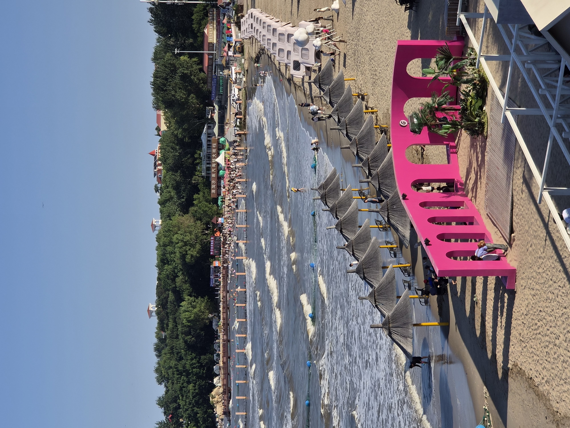 The image depicts a bustling beach scene in Qinhuangdao, China, likely in the popular Beidaihe district, known for its seaside resorts. The foreground is dominated by a vibrant pink, arched structure, resembling an entryway or stage, adorned with lush artificial foliage and palm trees. Behind this structure, rows of thatched-roof umbrellas with yellow poles provide shade for beachgoers, with some empty chairs visible beneath them.
Further back, the sandy beach transitions to the wavy ocean, where numerous people are enjoying the water, swimming, and wading. Safety buoys and nets delineate swimming areas, and a wooden pier extends into the sea, lined with more beach umbrellas and what appear to be small, tent-like structures. In the background, a dense line of green trees separates the beach from buildings and structures on a higher elevation. The sky is clear and blue, indicating a sunny day.
Several individuals are visible throughout the scene. On the beach, people are walking, playing, and tending to the area around the pink structure. In the water, swimmers and waders are scattered across the designated zones. The overall impression is one of a lively and popular tourist destination on a summer day.