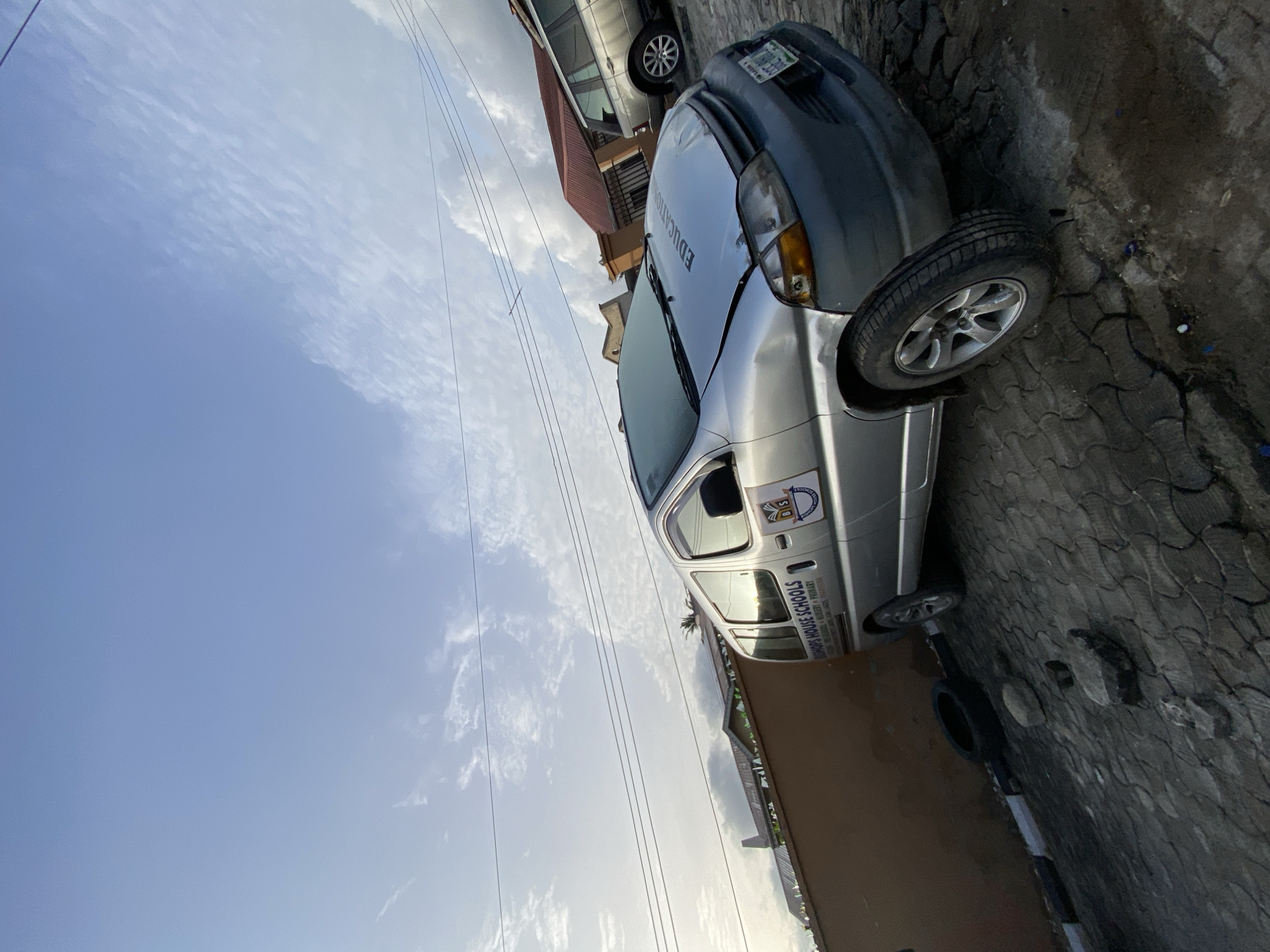 The image depicts two cars parked on a paved surface in Ado, Nigeria. One is a silver minivan with the words "EDUCATION" on the hood and "BISHOPS HOUSE SCHOOLS" with additional details on the side. The other is a dark-colored sedan. The environment includes a building with a red roof in the background and utility lines overhead. The sky is visible with scattered clouds.
