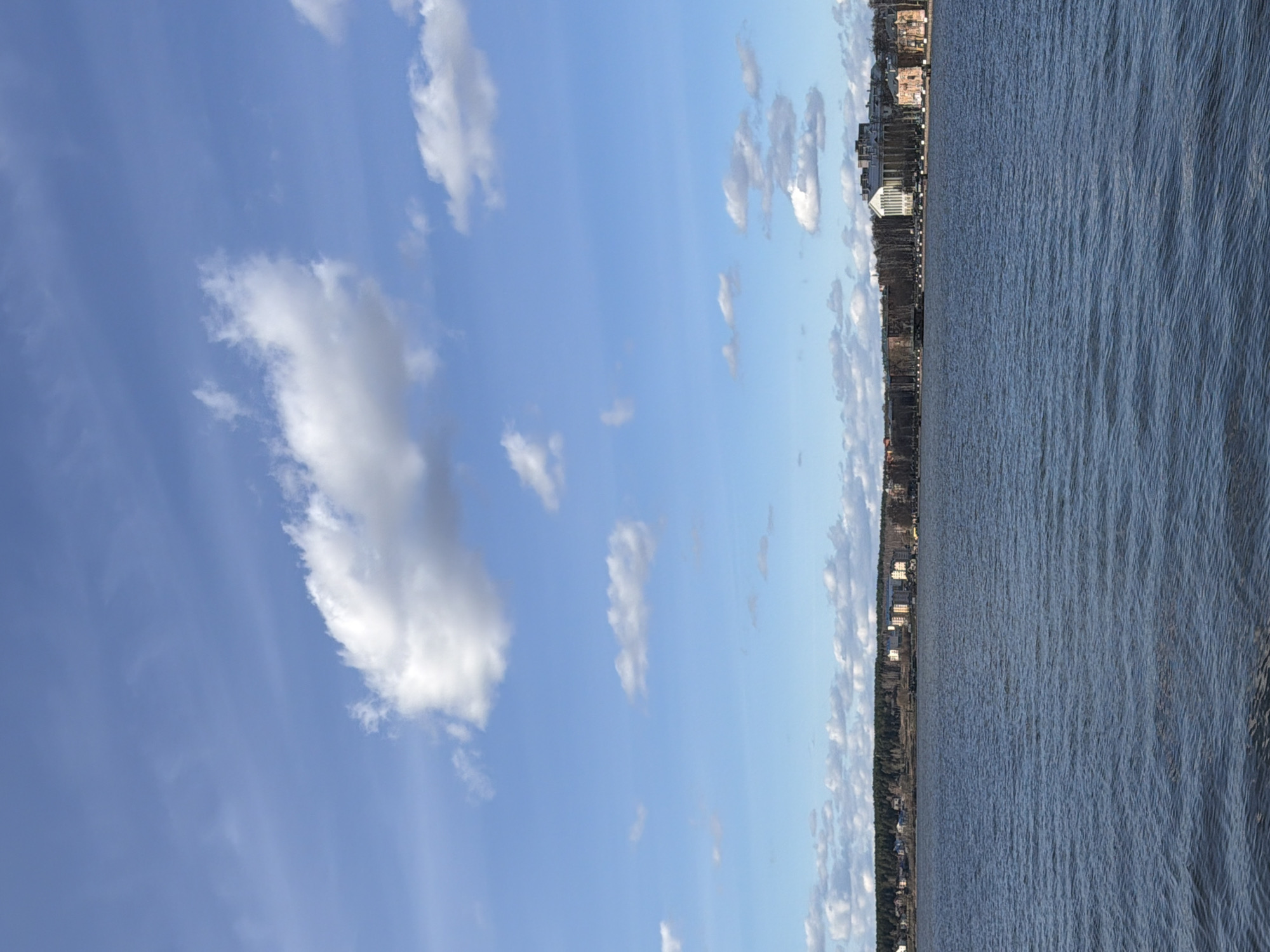 The image shows a body of water, likely a river or large lake, under a partly cloudy sky. The water surface is slightly rippled. Along the distant shoreline, there's a row of buildings and trees, suggesting an urban or suburban setting. Based on the context of the location, the image is likely captured at the Votkinsk Reservoir, in the city of Votkinsk, Russia. There are no visible people or specific actions occurring.