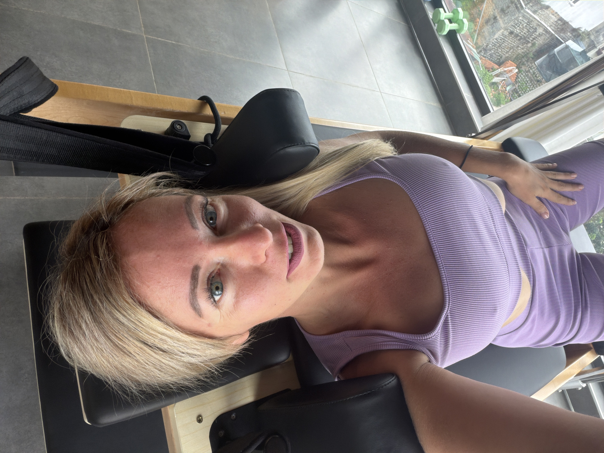 A woman with blonde hair and green eyes is lying down on a pilates reformer. She is wearing a purple ribbed tank top and matching leggings. Her left hand is resting on her stomach, and her right hand is visible. In the background, a window shows a view of rooftops and greenery. The pilates reformer is made of wood and black padding. There are also two green dumbbells visible on the windowsill. The flooring appears to be grey tile.