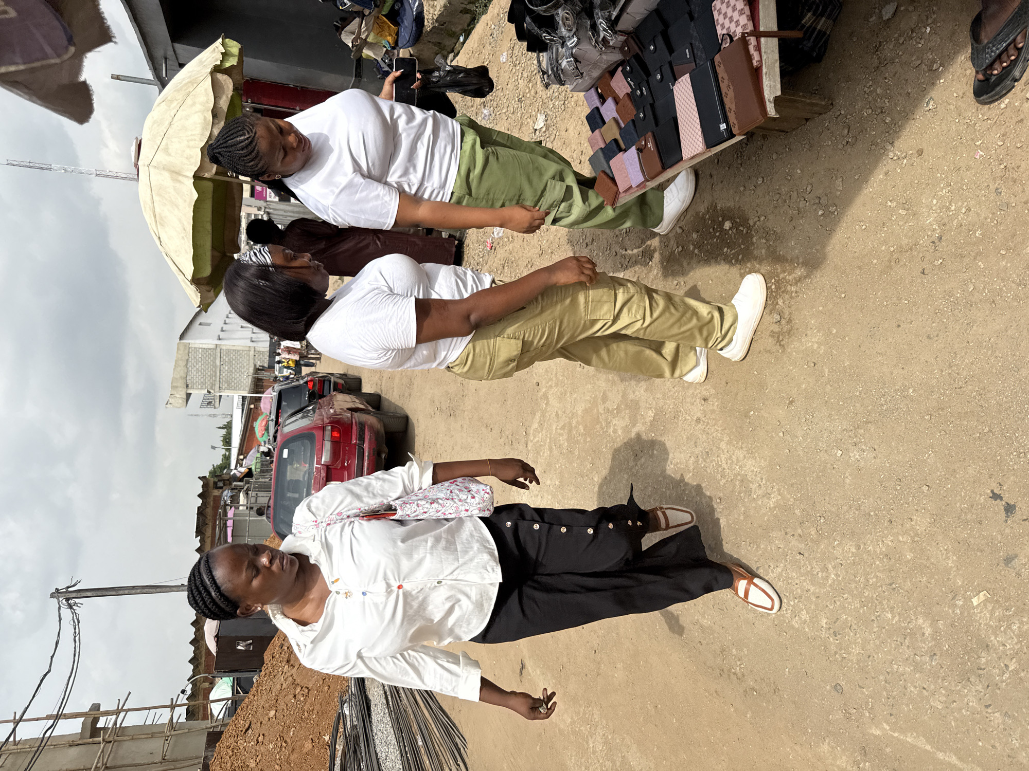 The image shows an outdoor scene in what appears to be a street market in Abuja, Nigeria. A woman in a white shirt and black pants walks on a dusty ground, with metal rods visible near her. Two women, wearing white shirts and khaki pants, stand near a stall displaying various wallets and bags. An umbrella provides shade for the stall. A red car is parked nearby. The sky is overcast.