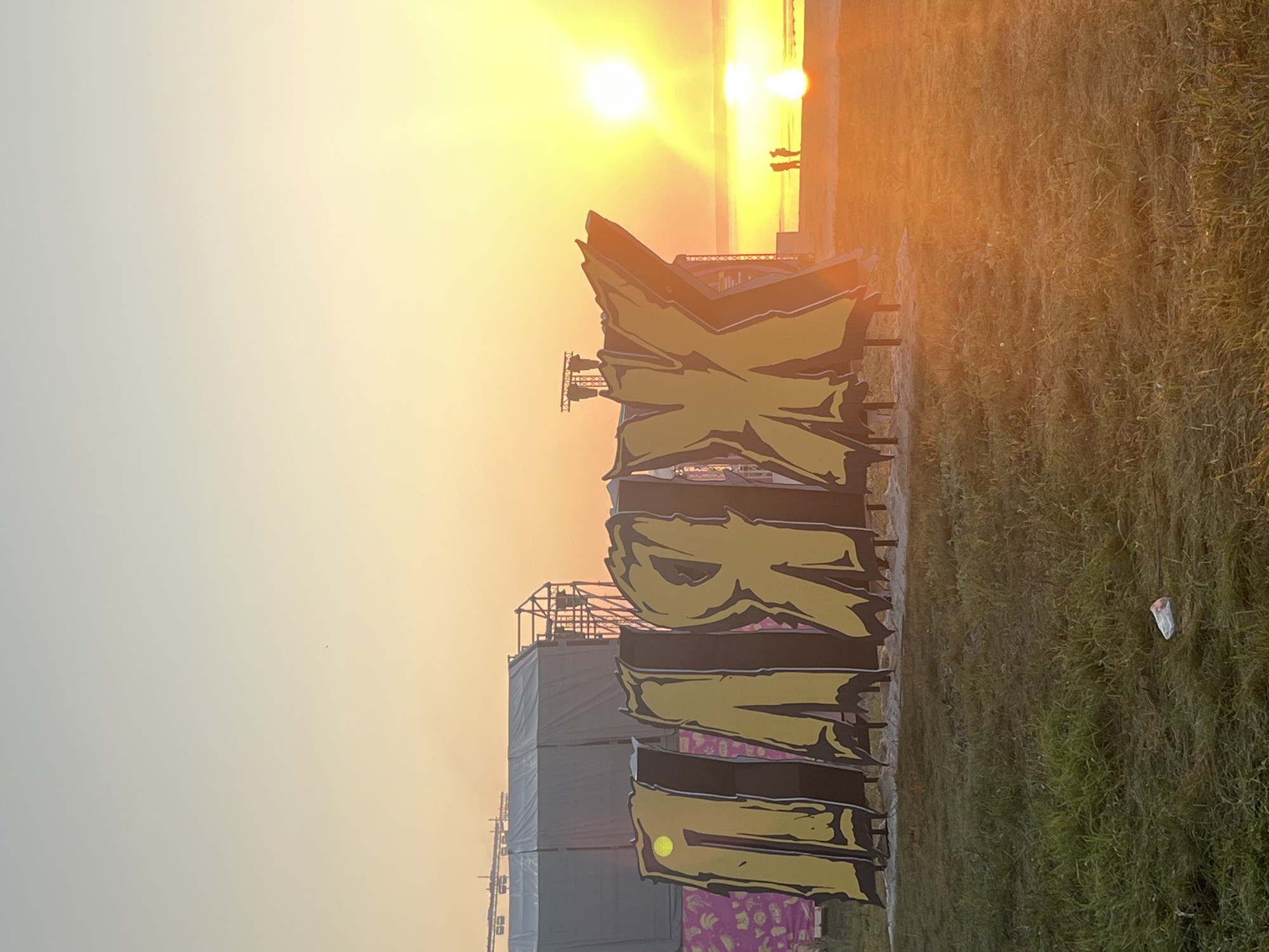 The image depicts a sunset over a body of water, likely a sea or large lake, viewed from a grassy area. In the foreground, large, stylized yellow letters with black outlines spell out "ПЛЯЖ" (which translates to "BEACH" in Russian). Behind the letters, a stage setup with tents and scaffolding is visible. Two figures are silhouetted on the beach in the middle distance, facing the setting sun. The sun is prominently featured, low on the horizon and casting a warm, golden light across the scene, creating lens flare and reflections on the water. The sky transitions from a pale blue at the top to a vibrant orange and yellow near the horizon. There is scattered litter on the grass in the foreground. The overall impression is an outdoor event or festival taking place at sunset on a beach. The location is specified as Surakovo, Russia.