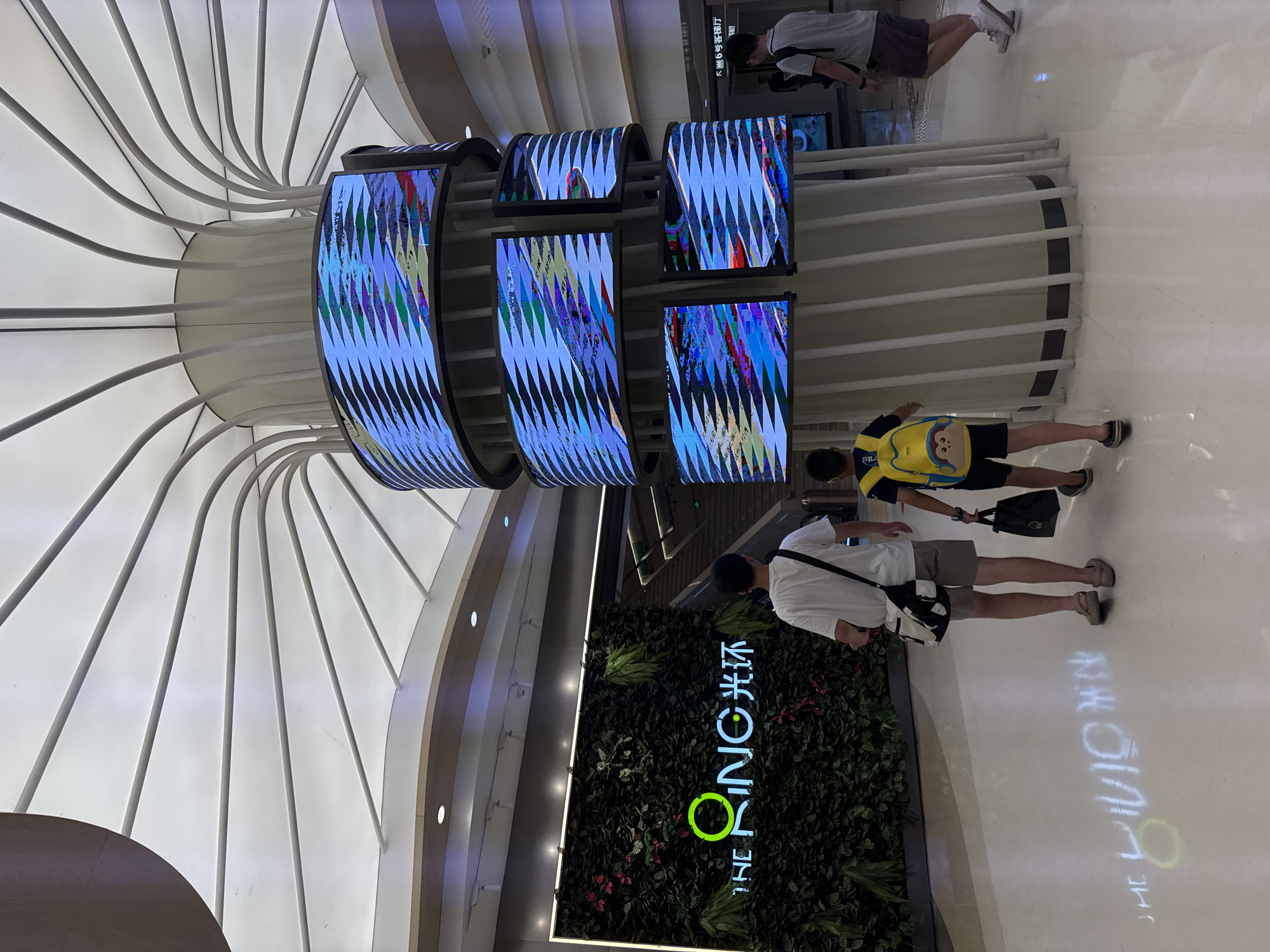 The image captures an indoor scene, likely a shopping mall or public space in Liangjiang, China. In the foreground, two individuals are walking away from the camera: an adult male on the left wearing a white t-shirt, khaki shorts, and carrying a crossbody bag, and a younger male on the right with a yellow backpack adorned with a monkey face, wearing a dark t-shirt and dark shorts. The floor is a light-colored, polished material, reflecting light and a projected graphic of "THE RING" with a circular logo and Chinese characters that translate to "Guanghuan" (which means ring or halo).
Dominating the background is a large, cylindrical structure with a distinctive ceiling resembling an inverted flower or umbrella, constructed with white fabric and supported by numerous thin, curved beams radiating outwards. Wrapped around the cylindrical structure are several curved LED screens displaying abstract, fragmented patterns of blue, white, and red. To the left of the pillar, a vertical wall is covered in lush green foliage, interspersed with red and green leaves, featuring a neon sign that reads "THE RING" with the same circular logo and Chinese characters. Stairs are visible in the mid-ground to the right. Other individuals are visible in the background, suggesting a public gathering space. The overall atmosphere appears modern and commercial.