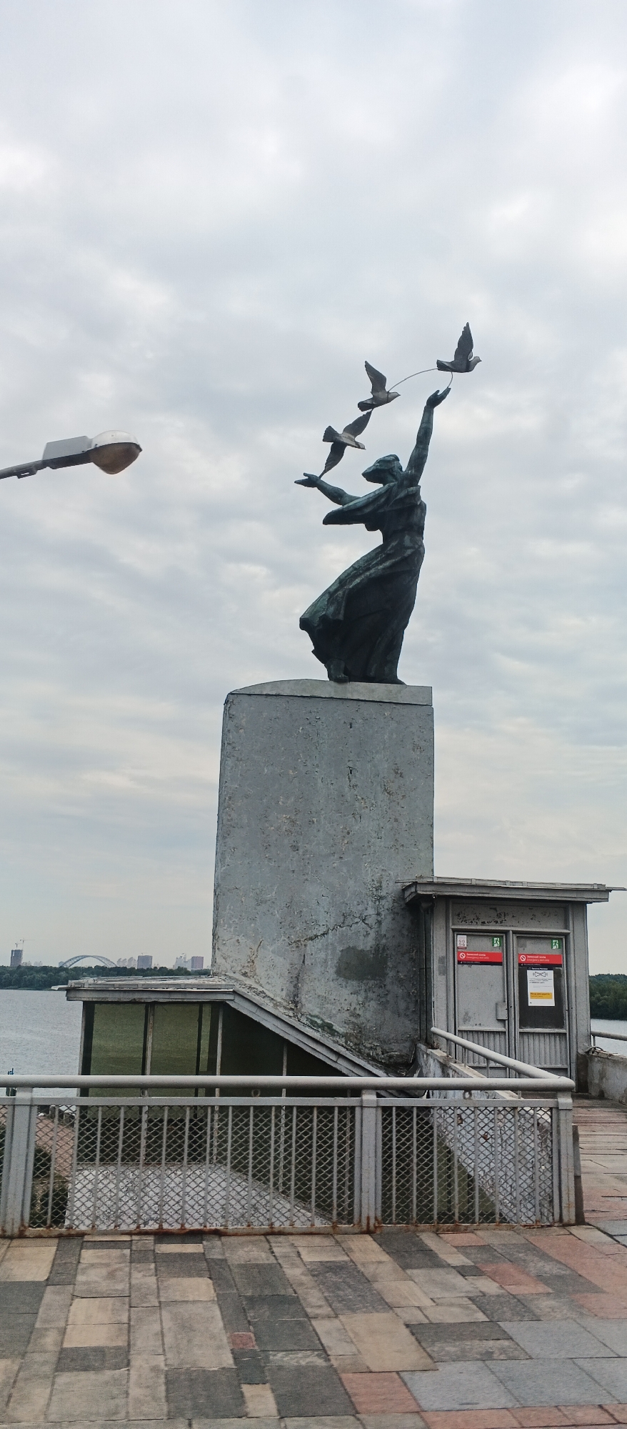 A bronze statue of a woman releasing three doves stands atop a concrete pedestal in Kyiv, Ukraine. The woman, depicted with her arms outstretched and her body leaning back, appears to be in motion. The doves are shown in flight, with one in the foreground and two slightly behind it. To the left of the statue, a street lamp extends horizontally into the frame. A small, gray building with a metal door and several signs is situated to the right of the pedestal. In the background, a body of water, a bridge, and city buildings are visible under an overcast sky. The foreground features a paved walkway with a metal railing.