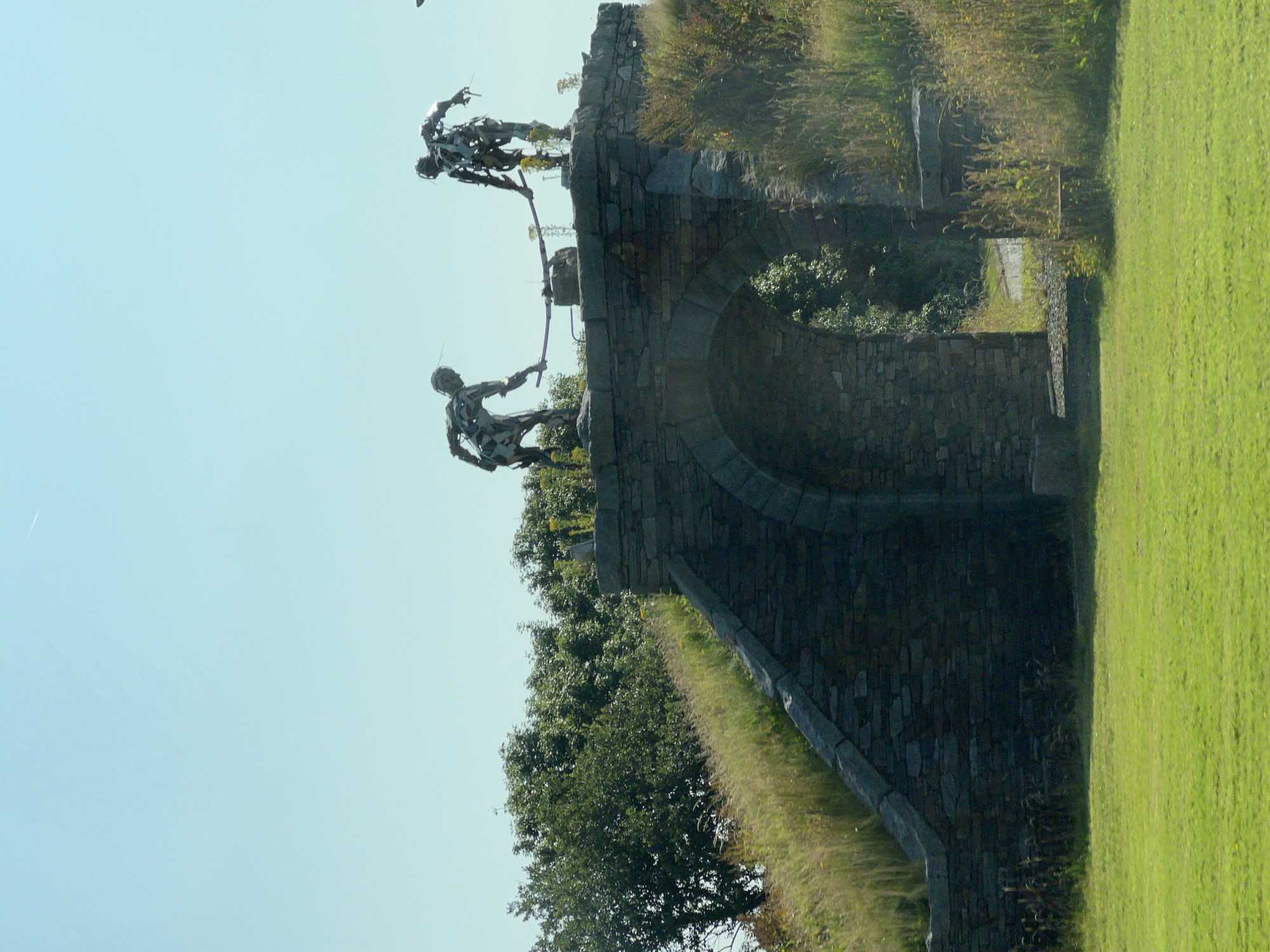 This is a medium shot, taken from a slight low angle, of the "Stone Arch and Figures" sculpture in Letterkenny, Ireland. The sculpture depicts two abstract figures made of welded metal, in a stooped posture, appearing to be holding a large beam or pole between them. They are positioned atop a stone archway, which is constructed from rough-hewn grey stones. The archway is surrounded by grassy slopes covered in tall, dry grasses and wildflowers, with a dense green tree line in the background. The sky is a clear, pale blue with a faint contrail visible. The foreground features a well-maintained green lawn. The sculpture's figures are dark and metallic, contrasting with the natural, earthy tones of the stone and vegetation. The overall impression is one of an outdoor public art installation integrated into a landscaped environment.
