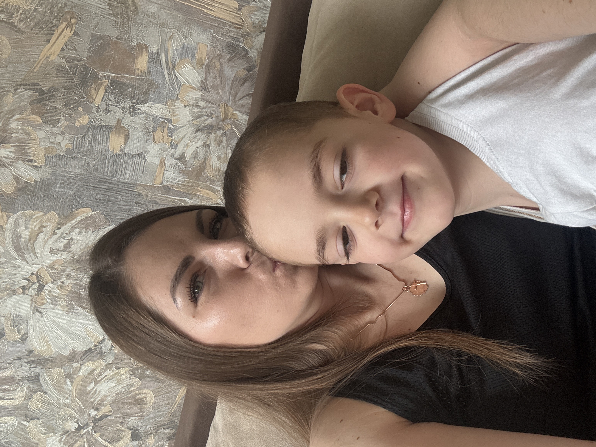 A woman and a child are lying down in what appears to be an indoor setting. The woman has brown hair and is wearing a black top and a necklace. The child, who seems to be a young boy, is wearing a white tank top. The background shows a wall with a textured floral pattern. The location is Раздолье, Russia. There are no other visible people or objects. The woman appears to be kissing the child on the cheek.