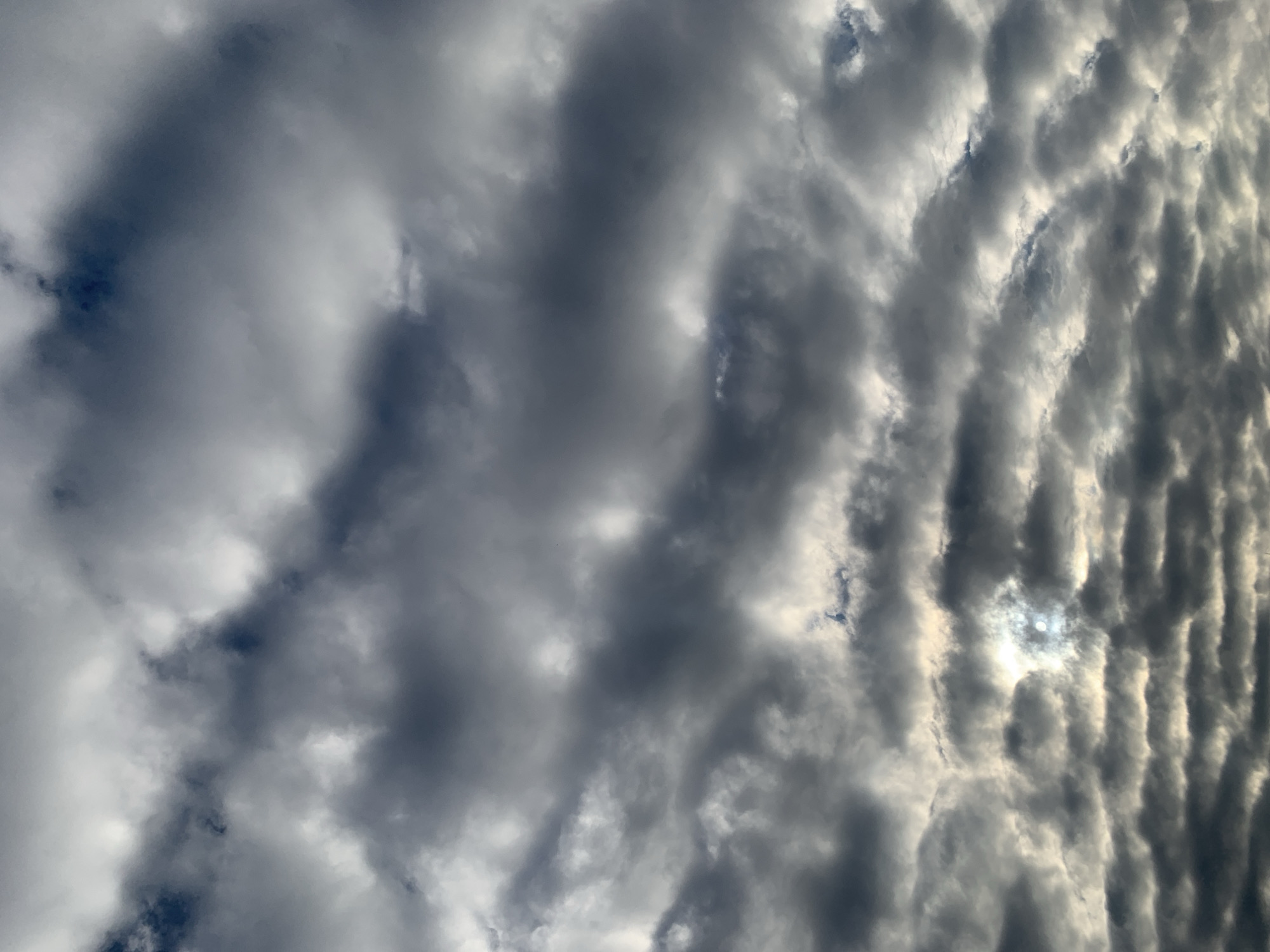 This image depicts a cloudy sky with the sun partially visible, creating a luminous effect. The clouds are varied in texture and shade, ranging from dark gray to lighter, wispy formations. The sun's rays pierce through some of the denser cloud cover, illuminating parts of the sky with a bright, diffused light. The overall impression is one of dynamic atmospheric conditions. The scene is a sky view and does not contain any elements that would indicate a specific location within the city of Kolomyia, Ukraine.