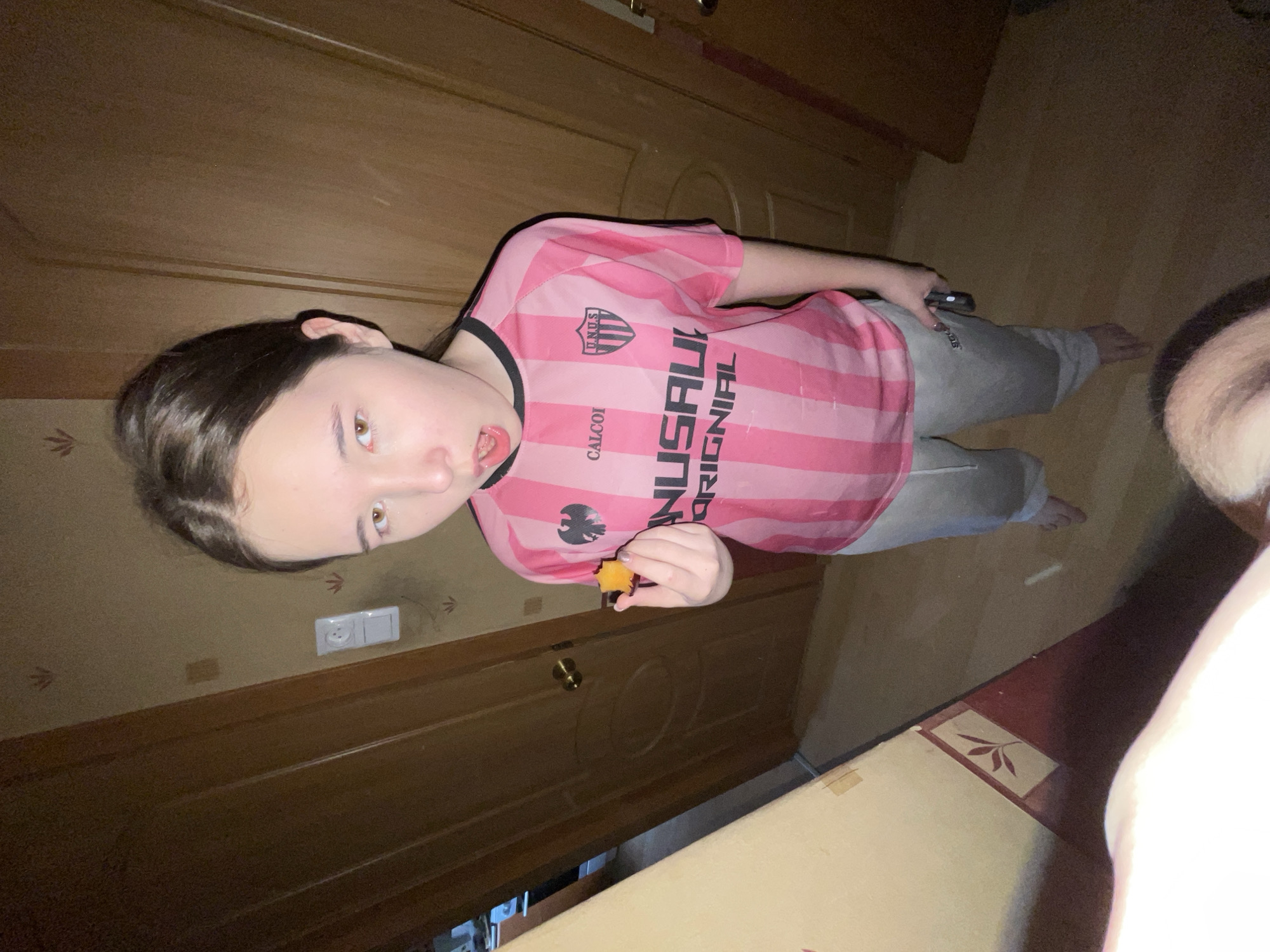 A young person is indoors, in what appears to be a room with wooden paneling and doors. The person is wearing a pink and black striped jersey with the word "NUSALI" on the front and grey sweatpants. They are holding a small orange object, possibly a piece of food, near their mouth. The location cannot be verified as being in Saint Petersburg, Russia, as there is no indication of that in the photograph.