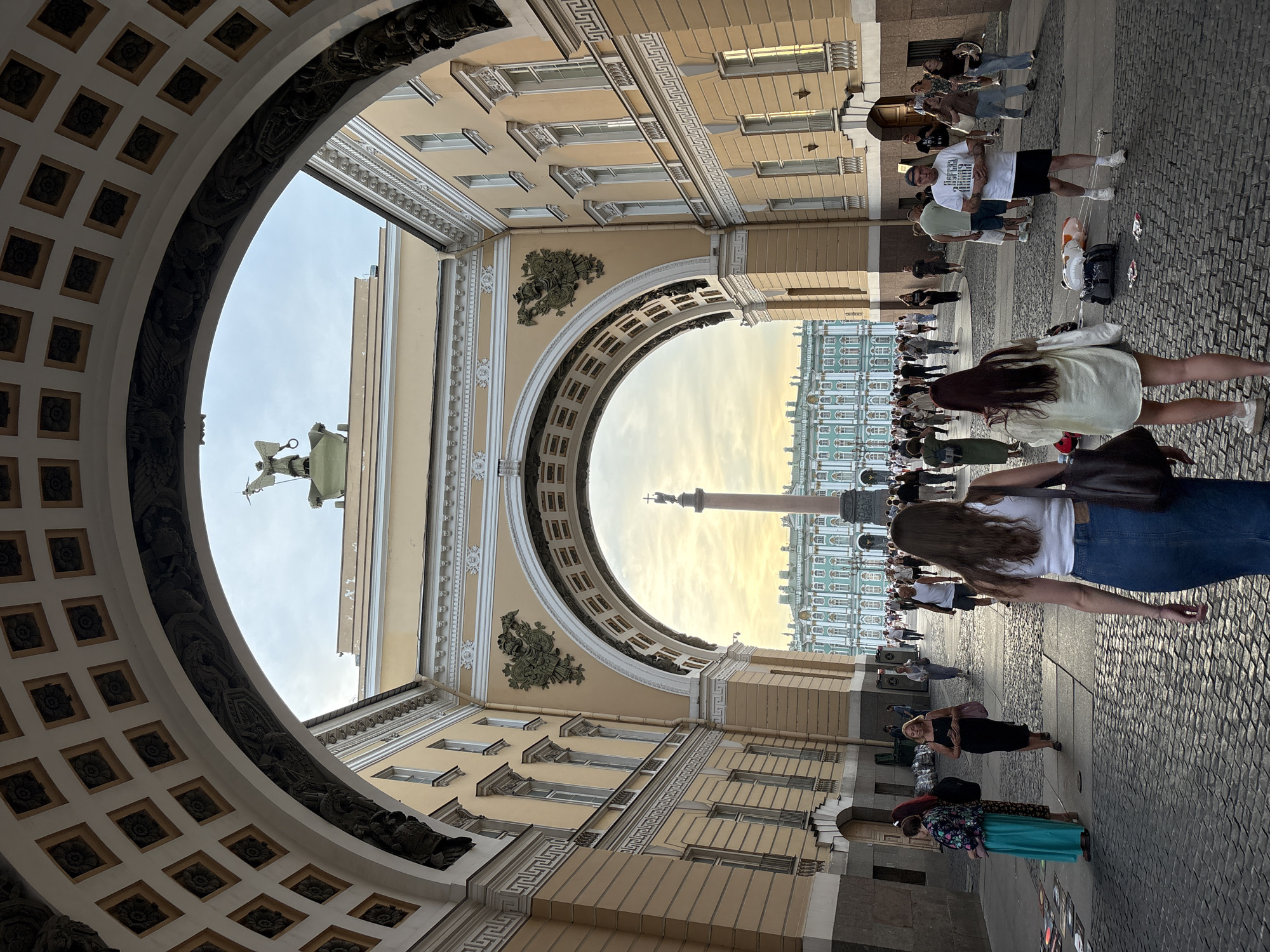 The image captures a view through a grand triumphal archway in Saint Petersburg, Russia, leading to Palace Square. The arch, part of the General Staff Building, is ornately decorated with bas-reliefs and a patterned ceiling. Beyond the arch, the expansive Palace Square is visible, dominated by the majestic Winter Palace with its turquoise and white facade. In the center of the square stands the Alexander Column, a towering granite monument topped with an angel.
The foreground is a cobblestone plaza where a diverse group of people are walking, standing, and interacting. Many are tourists, evident from their casual attire and the presence of backpacks. Two women in the immediate foreground are walking away from the camera, one wearing a denim skirt and white tank top, the other in a light-colored oversized shirt and shorts. To their left, two women in long dresses are standing, one in a bright turquoise maxi dress, and the other in a patterned dress. Further into the square, more individuals are dispersed, some in small groups, others walking alone, navigating the large open space. Several balloons and bags are scattered on the ground near a man in a white t-shirt and black shorts, suggesting a possible street performance or a temporary setup. The sky above is overcast with hints of golden light, indicating either early morning or late afternoon.