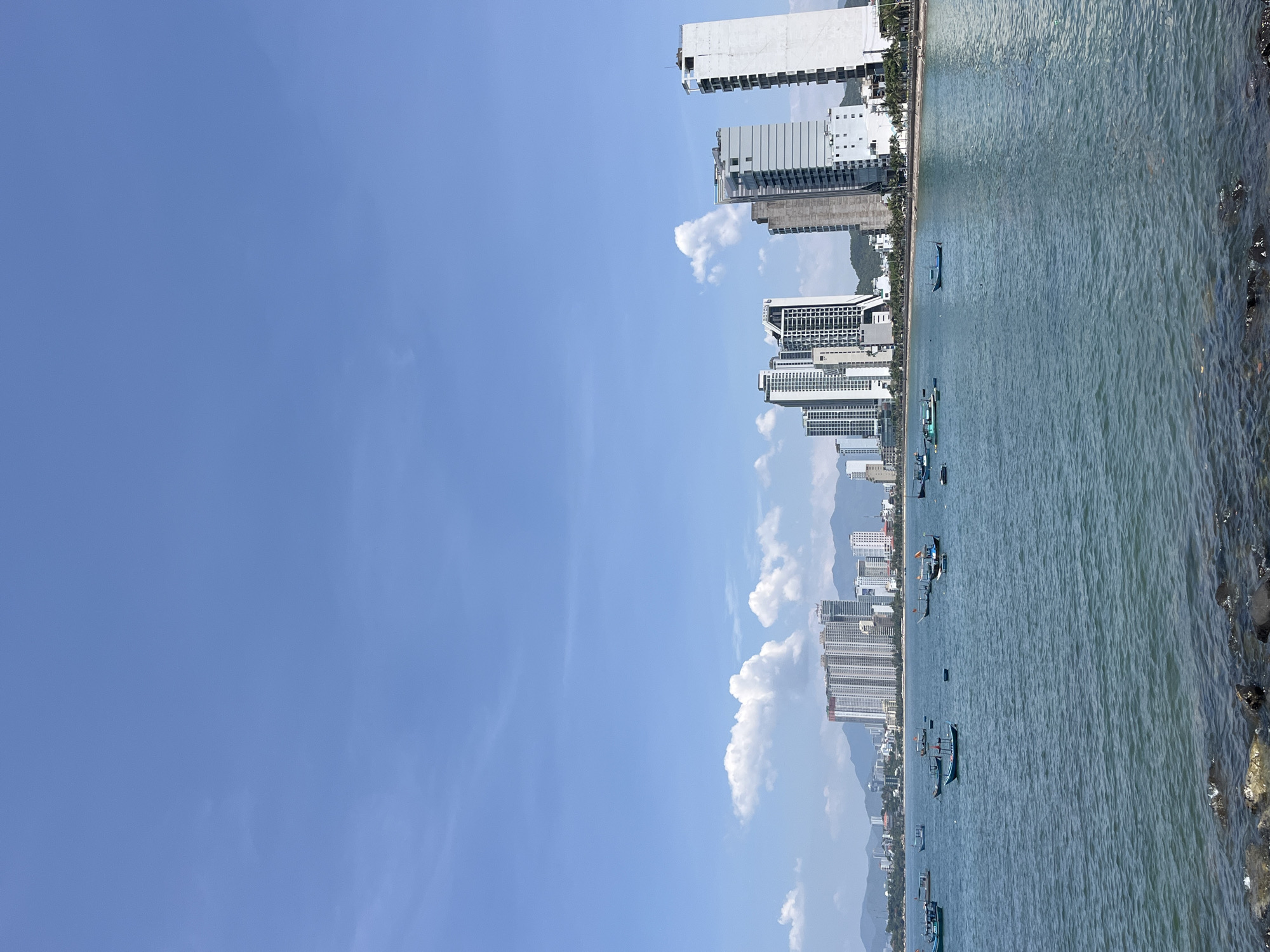 This image captures a wide-angle view of the coastline of Bac Nha Trang Ward, Vietnam, on a clear, sunny day. The upper half of the frame is dominated by a bright blue sky with scattered white cumulus clouds.
In the mid-ground, a prominent urban skyline stretches horizontally across the scene. Numerous high-rise buildings, predominantly white and grey, line the waterfront. Some structures appear to be newly constructed or undergoing completion, indicated by exposed concrete and framing on their upper sections. Mountains or hills are visible in the background behind the city, partially obscured by the urban development. A paved road and a strip of vegetation, including palm trees, run along the base of the buildings at the immediate shoreline.
The lower half of the image features a calm body of water, likely a bay or the sea, with a blue-green hue and subtle surface ripples. Numerous small, traditional fishing boats, mostly painted in shades of blue and green, are scattered across the water, appearing to be anchored.
In the immediate foreground, a rocky shoreline is visible, consisting of dark, irregularly shaped stones and boulders partially submerged or wet from the water. No individuals are discernible within the frame.