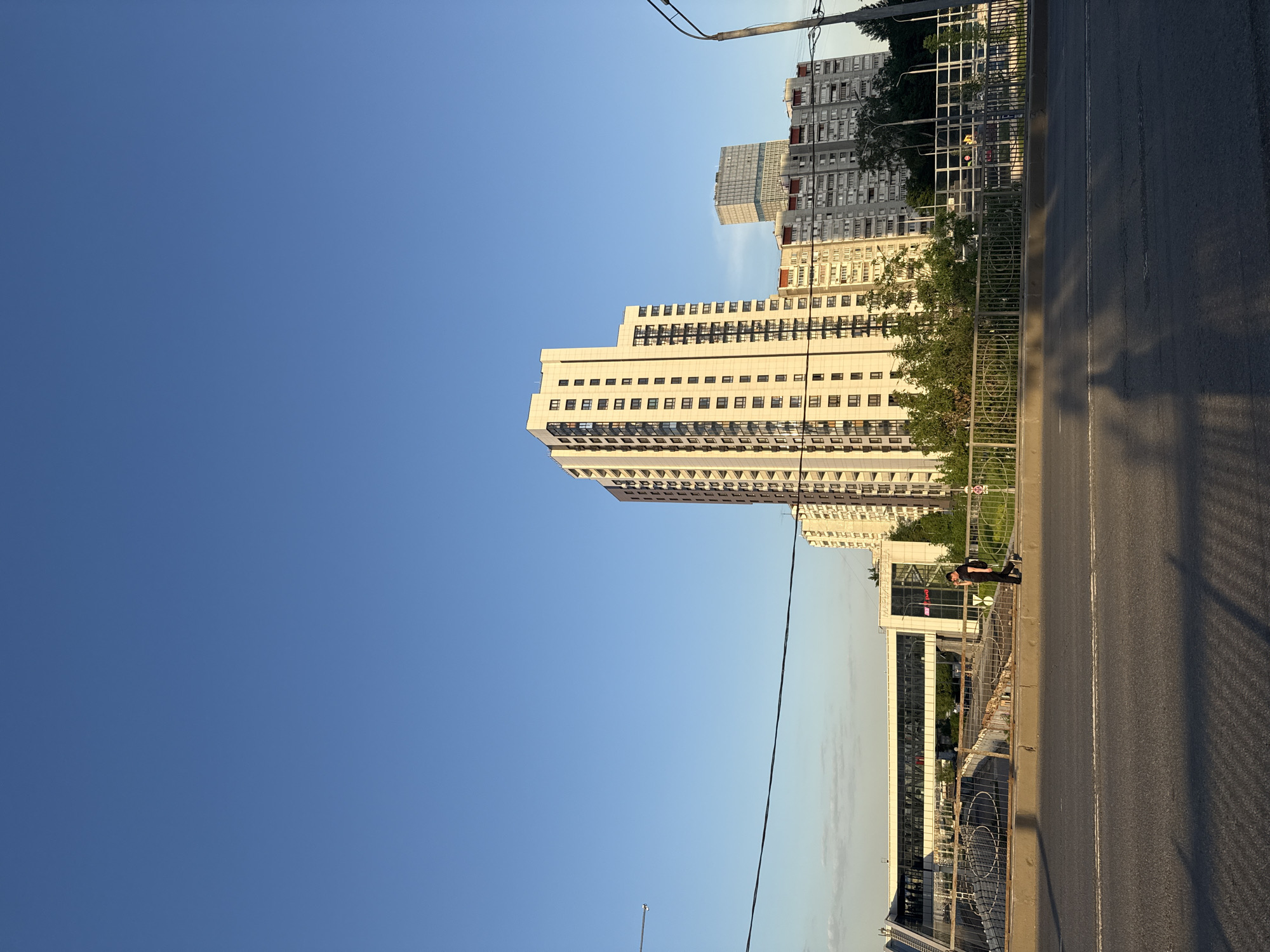 The image depicts a street view in Moscow, Russia, featuring modern architecture and urban infrastructure. On the right side of the frame, there's an asphalt road with painted white lines marking lanes. A metal railing runs along the roadside, separating the street from a lower level. A tall, light-colored building with numerous windows dominates the skyline in the background. To the left, the sky is a clear, deep blue. A power line stretches across the image, adding a detail of the urban environment. Additionally, there's a pedestrian visible walking along the bridge, suggesting the image captures a moment of city life.