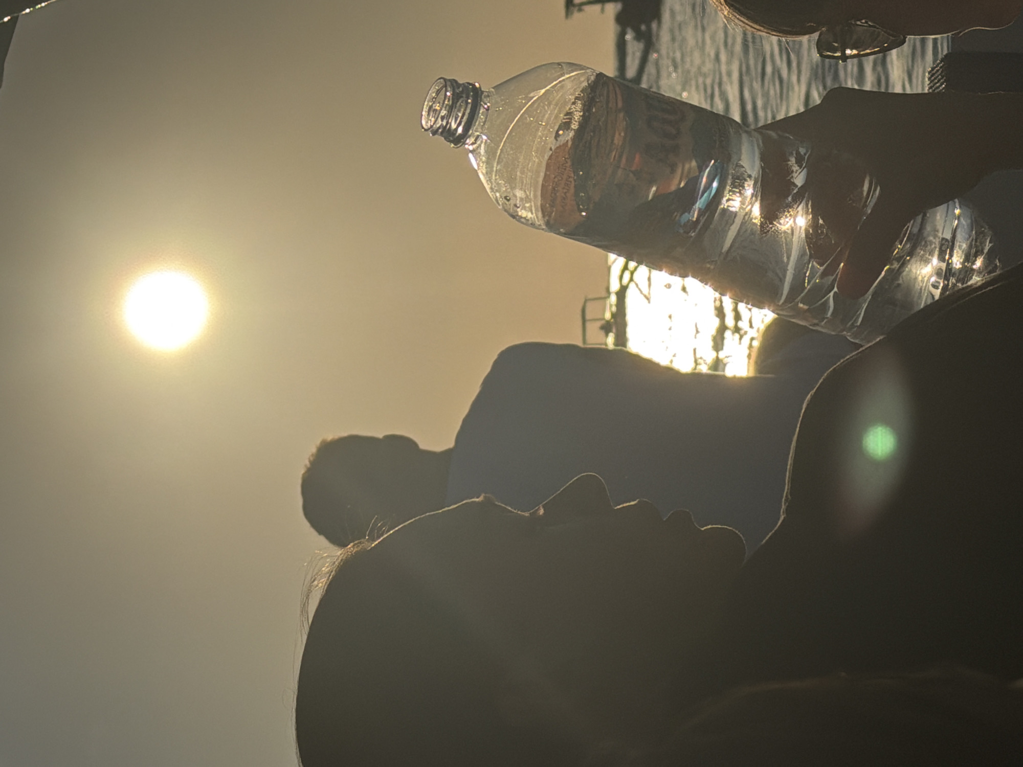 A low-angle, silhouette shot captures a sunset over the waters of Seririt, Indonesia. In the foreground, a person's profile is visible on the left, their features obscured by shadow. To the right, another person's hand holds up a clear plastic bottle of water, which catches the sunlight. In the background, two other figures stand silhouetted against the bright sky. The sun, a bright orb, is positioned high in the frame, casting a warm glow over the scene. The water reflects the sunlight, creating a shimmering effect. Small boats are visible in the distance on the water. The overall impression is one of a peaceful, late afternoon scene by the sea.
