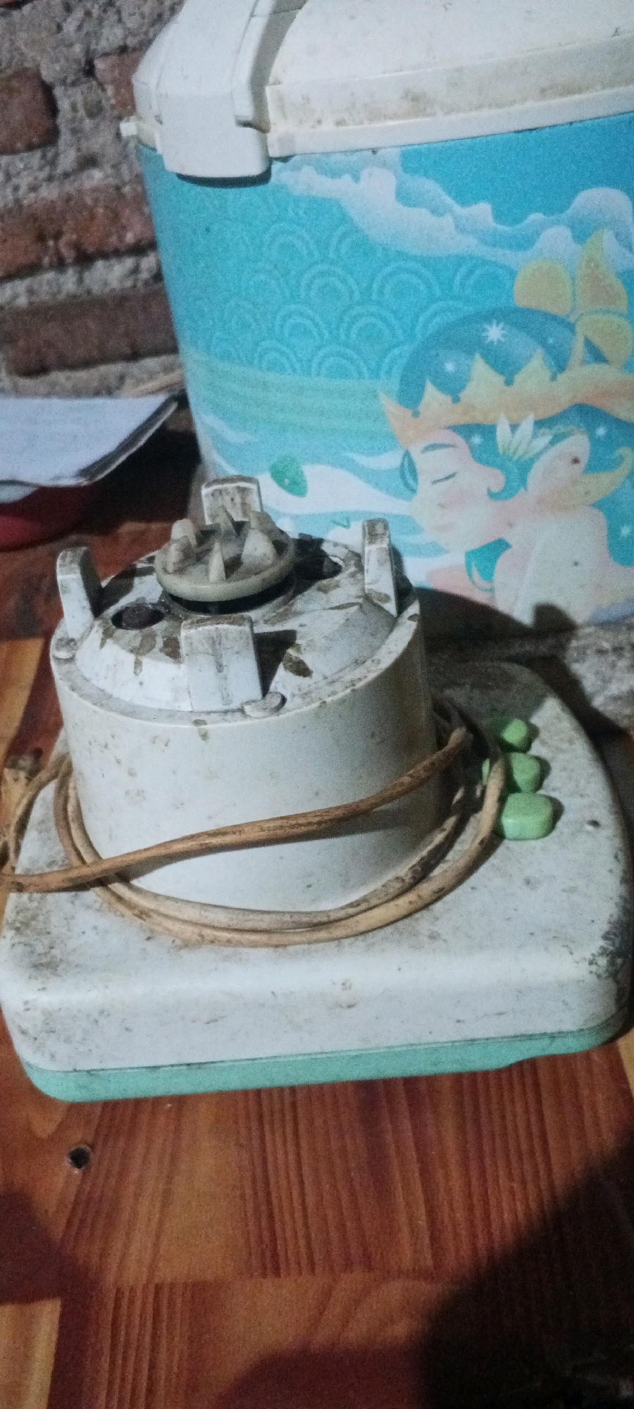 The image depicts a close-up, outdoor, eye-level shot of a blender base on a wooden surface. The blender base is a light-colored, rectangular object with a slightly elevated circular component on top, where the blades would attach. This component shows signs of wear and is somewhat dirty. A brown electrical cord is coiled around the base, and three small, rectangular green objects are placed on the right side of the base.
In the background, a portion of a blue container with a patterned design and an illustration of a person with a crown is visible. To the far left, a brick wall is present. The overall scene appears to be cluttered, with dust and dirt evident on the blender and the surrounding surfaces. The lighting suggests it is daytime. The image does not provide specific information about the city of Penanggungan, Indonesia.