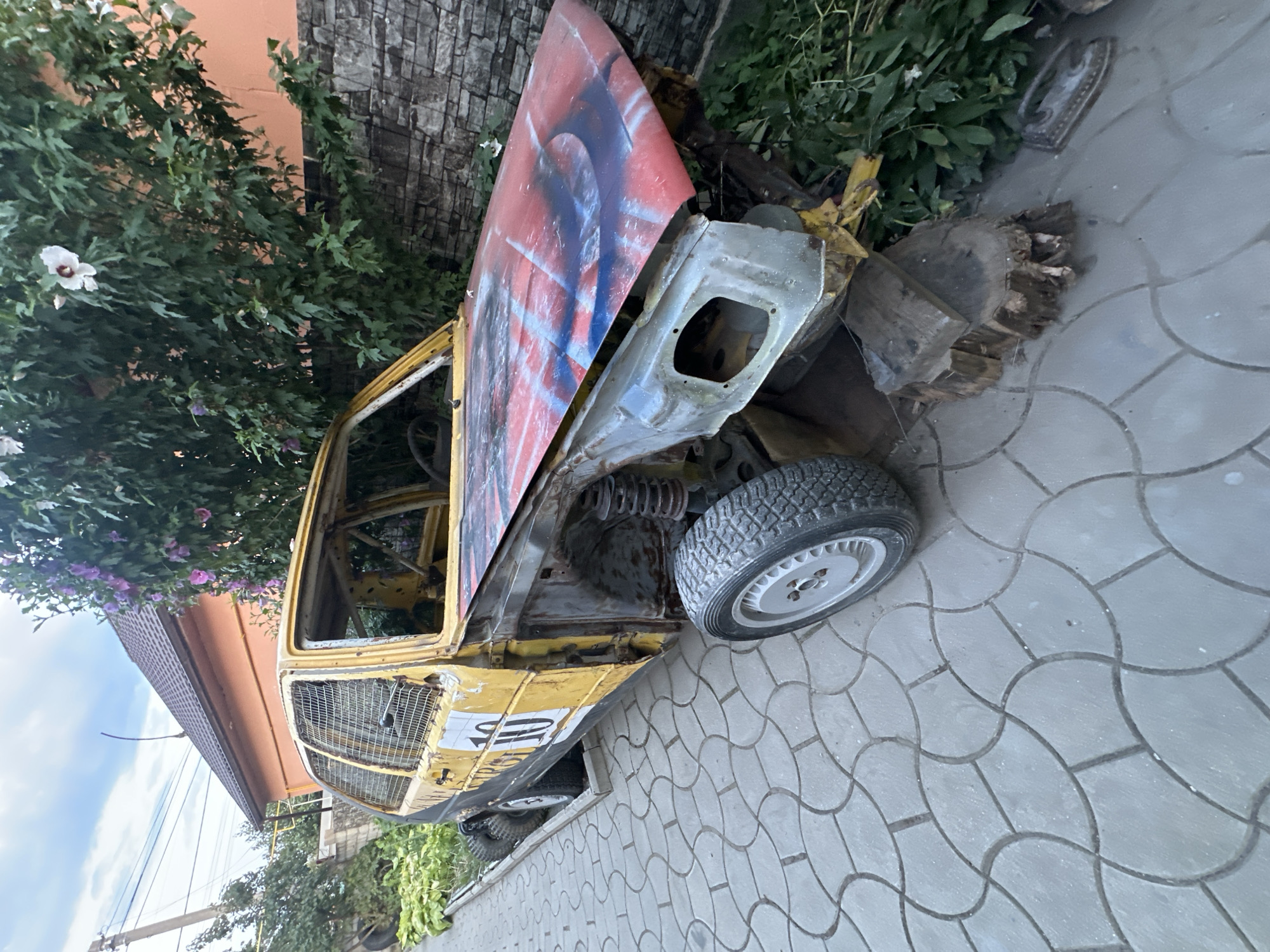 A yellow, heavily modified, stripped-down car, possibly a Volkswagen Golf, sits on a paved driveway in Comrat, Moldova. The car has a roll cage visible inside, the hood is painted with red and blue spray paint, and the body panels show significant rust and damage. It appears to be in a state of disrepair or as a race car preparation project. The car is surrounded by greenery, including a flowering bush, and a stone wall is visible in the background. A residential building with an orange facade and a tiled roof is also in the background. The scene is outdoors, under a cloudy sky.