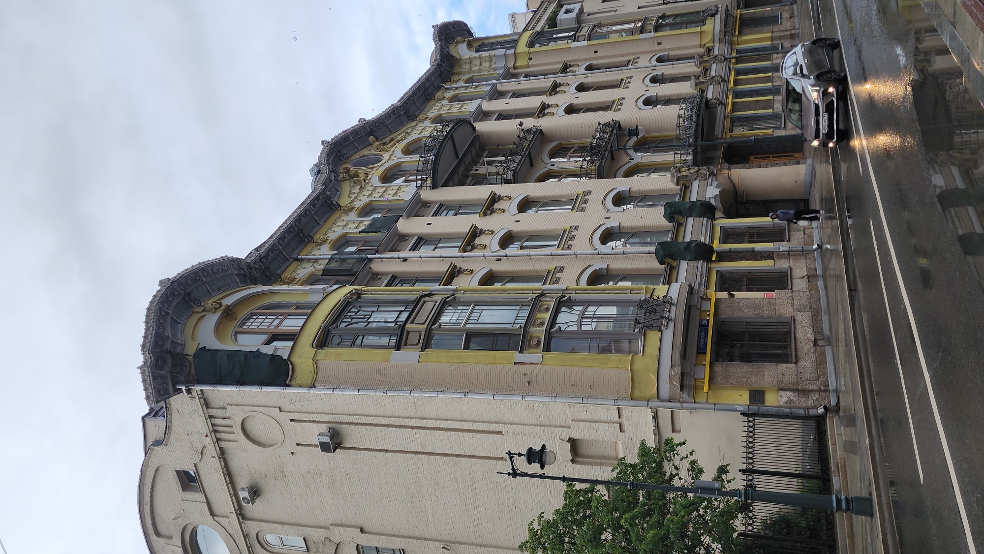 The image depicts a building in Moscow, Russia, identifiable by the street name "Кузнецкий", which is visible on a blue sign. The building's architectural style is ornate, featuring multiple levels with varied window designs, including arched and bay windows, some with decorative iron balconies. The facade is painted in light yellow and beige tones with darker yellow accents highlighting architectural details.
A car is visible on the wet, reflective street, and a person stands on the sidewalk near the building. Street lamps and some greenery (trees) are also present. The sky is overcast, suggesting rainy or cloudy weather.