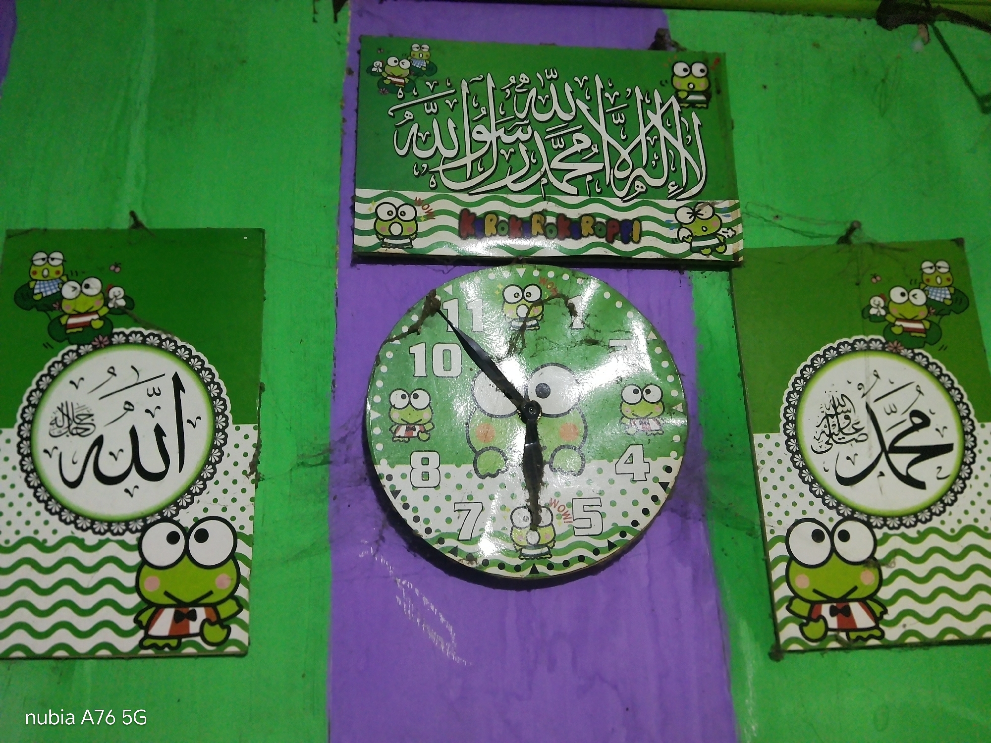 In Binuang, Indonesia, a wall is decorated with Islamic calligraphy and a clock, all featuring cartoon frogs. The green wall is adorned with three framed pieces. The central piece is a rectangular plaque with Arabic calligraphy in white script, bordered by a pattern of green and white wavy lines and smaller frog illustrations. Below this plaque, a circular clock with a green face, also adorned with frog characters and numerals, is mounted on a purple painted section of the wall. To the left and right of the clock, two square plaques, similar in design to the central plaque, display Arabic calligraphy within a circular frame and a cartoon frog at the bottom. The overall impression is a whimsical and religiously themed decoration in a residential setting.
