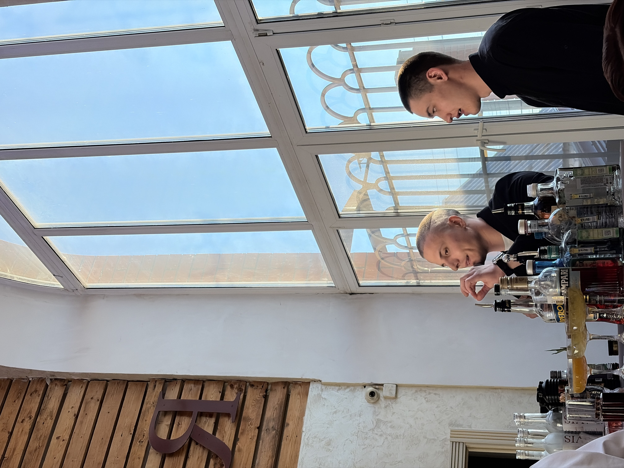 Two men are standing behind a bar filled with bottles of liquor. The man on the left is reaching toward a bottle, while the man on the right is looking on. The bar is in a room with a glass ceiling that lets in natural light. There is a wooden wall with a metal letter "R" on it. A security camera is visible on the wall. The image was taken in Neftekamsk, Russia.
