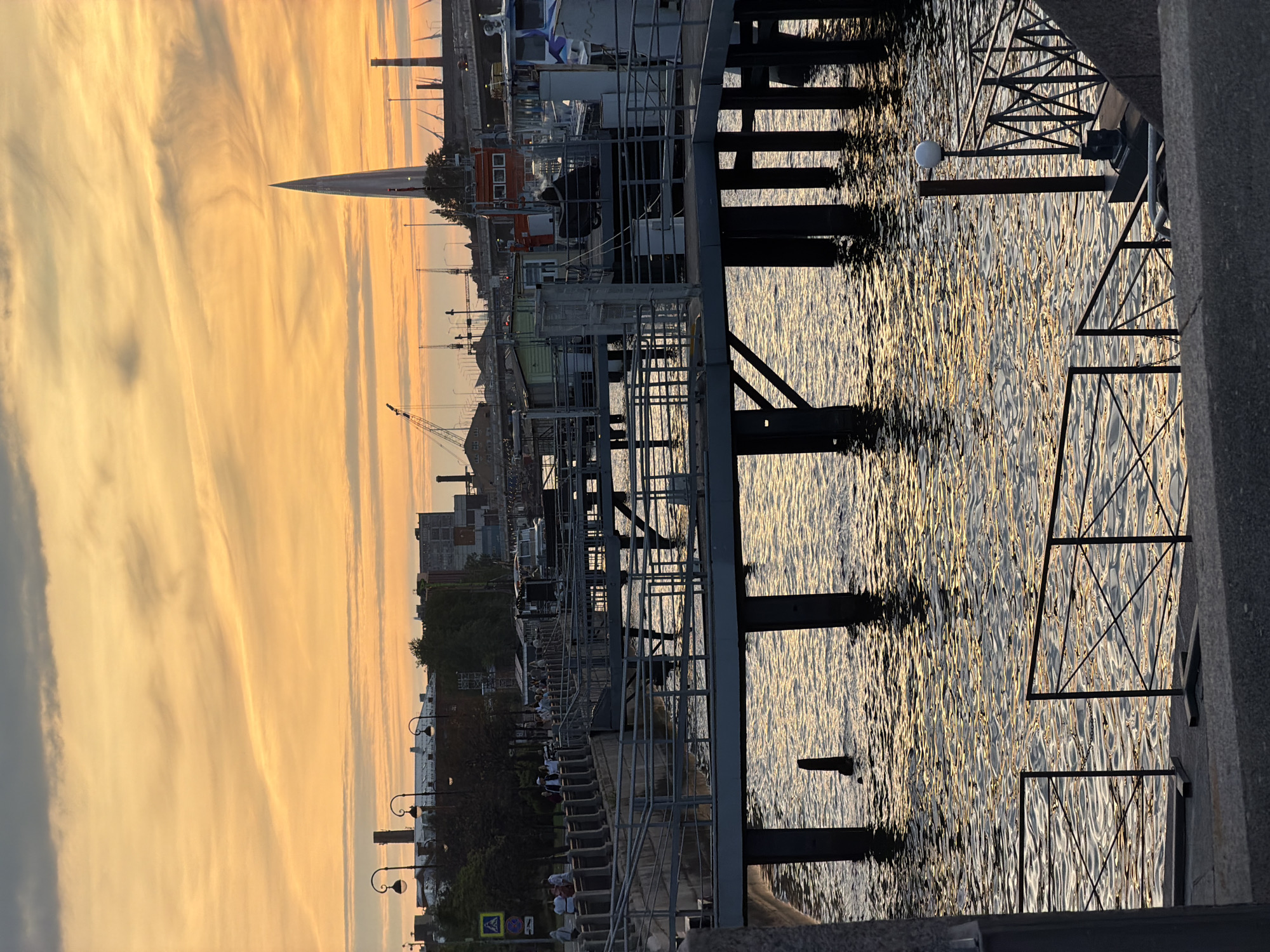 The image depicts a sunset over a waterfront area in Saint Petersburg, Russia. The sky is filled with dramatic orange and yellow clouds, casting a warm glow on the scene. In the foreground, there is a concrete embankment with metal railings. A series of piers extend into the dark water, supported by dark pilings. Boats are docked at the piers, and some people are visible walking along the waterfront. In the background, industrial buildings, cranes, and a distinctive modern skyscraper with a pointed spire are silhouetted against the sky. The water reflects the colors of the sunset, creating a shimmering effect.