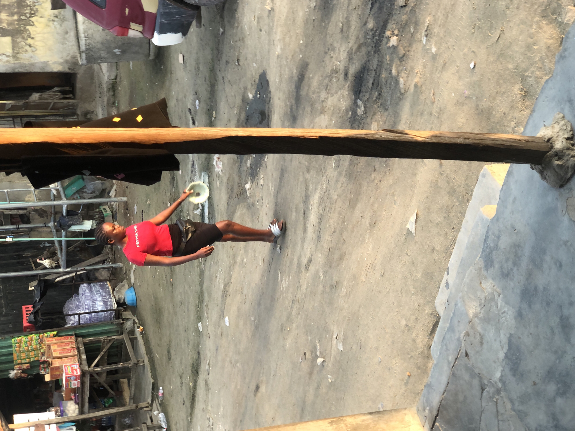 The image depicts an outdoor scene in what appears to be a commercial or residential area within Ado, Nigeria, under direct sunlight, suggesting late afternoon or early morning. A young Black female is centrally positioned, walking from left to right. She wears a red short-sleeved t-shirt with "PEPPER THE" printed in white, a black short skirt or shorts, a black fanny pack, and silver-and-white slide sandals. She holds a light-colored, shallow bowl in her right hand, extended downwards. Her hair is styled in braids.
In the immediate foreground on the right, a vertical, square-profile wooden post, dark brown with sunlit highlights, rises from a concrete base. To the left foreground, a section of a grey concrete step or ledge is visible.
The ground across the scene is unpaved, consisting of packed dirt or sand, scattered with small pieces of debris and litter.
In the background on the left, a stall or shop with a green corrugated metal facade is visible. It features a wooden display table holding multiple red-and-white cardboard boxes, likely containing food items. Various packaged goods are hung from the green wall. Behind the table, bundles of plastic-bottled water are stacked. A blue plastic basin rests on the ground near the stall. To the right of the stall, a white chicken is perched on a metal structure.
Further to the right in the background, a worn building with a light-colored concrete wall and an open doorway is visible. A red vehicle, specifically the rear bumper (partially painted white and dark) and a tire, is parked on the right side of the frame. Behind the wooden post, a black garment adorned with yellow diamond patterns hangs on an unseen support.