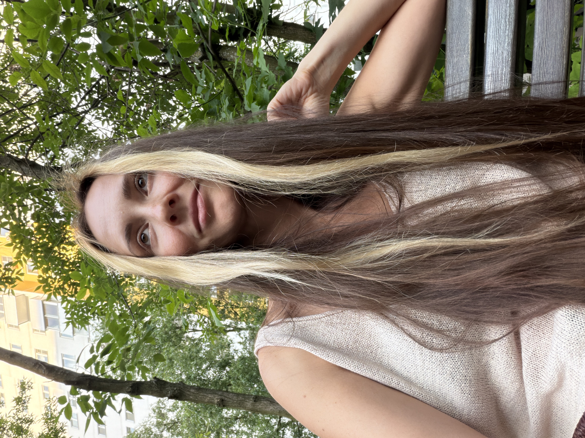 A woman with long, multi-toned brown and blonde hair sits on a wooden park bench, looking upwards and to her right. She is wearing a light-colored, knitted tank top. Her left arm is bent, with her hand resting near her head and her right arm is relaxed at her side. Behind her, lush green trees and foliage are visible, with a tall, light-colored building in the background, suggesting an urban park setting. The lighting indicates it is likely daytime.