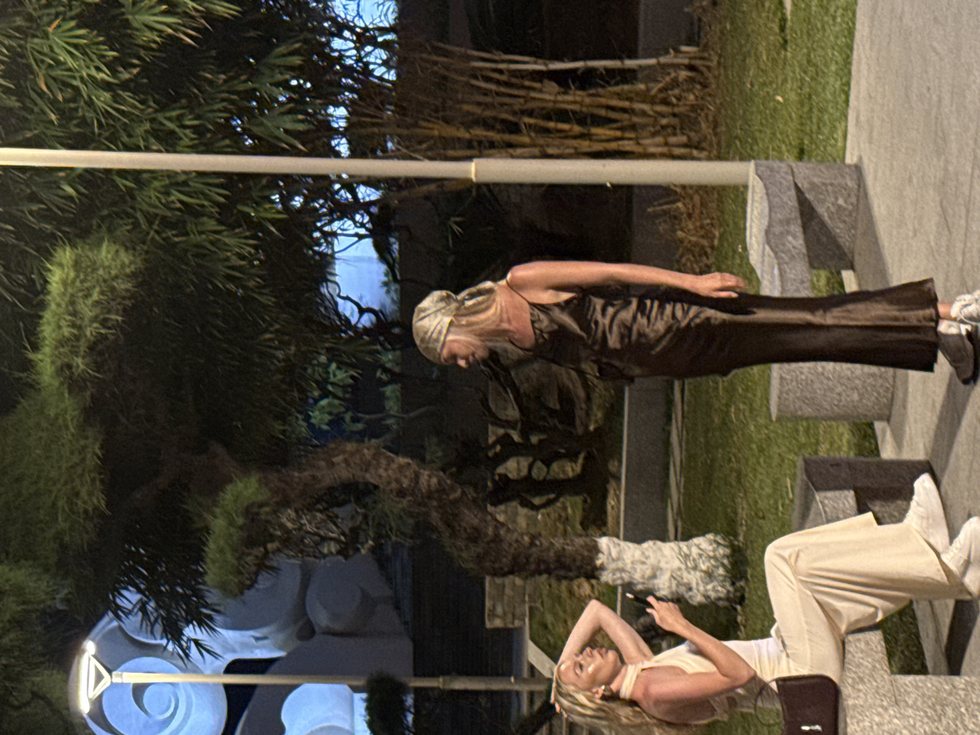 The image shows two women in an outdoor urban setting, likely at night in Nha Trang Ward, Vietnam. The woman standing wears a dark brown dress and white shoes. The woman lying down wears white pants and a tank top. The location has a modern design, with stone benches and possibly illuminated decorations. There are trees or large plants visible in the background, and possibly some bamboo.
