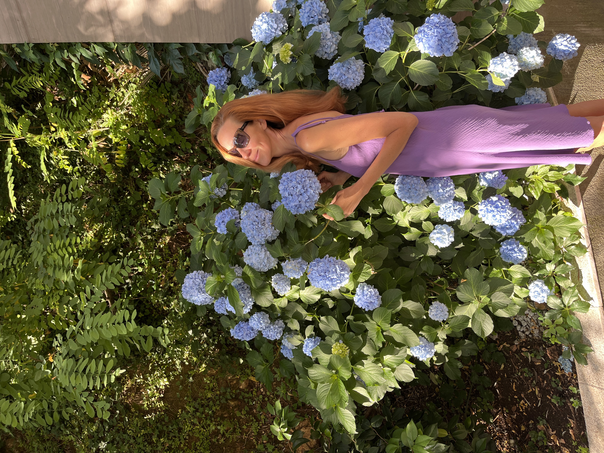 A red-haired woman wearing sunglasses and a purple dress stands beside a large bush of blue hydrangeas. She is looking up and to her left, with her right hand holding a hydrangea bloom. The background consists of lush green foliage and a textured wall. The scene appears to be outdoors in a garden setting, likely in the city of Budva, Montenegro, given the characteristic Mediterranean vegetation. The lighting suggests it is daytime, with sunlight casting shadows on the wall.