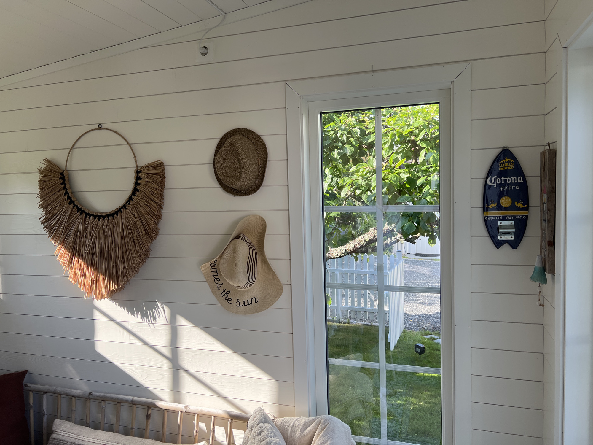 The image shows an interior view of a room with white horizontal shiplap walls. On the left wall, there is a large, circular, woven straw wall hanging with fringe. Below that, a brown woven hat hangs on the wall, followed by a larger, wide-brimmed straw hat with black ribbon trim, inscribed with "comes the sun." A wooden bench with a light-colored cushion is visible at the bottom of the frame. A window on the right wall looks out onto a green lawn, a white picket fence, and trees. On the wall to the right of the window, a blue surfboard-shaped sign with "Corona Extra" printed on it is mounted, along with a bottle opener. A small blue and white bell hangs below the sign. The scene appears to be a beach-themed interior, possibly a sunroom or beach house, located in Larkollen, Norway, a coastal town known for its beaches and summer atmosphere.