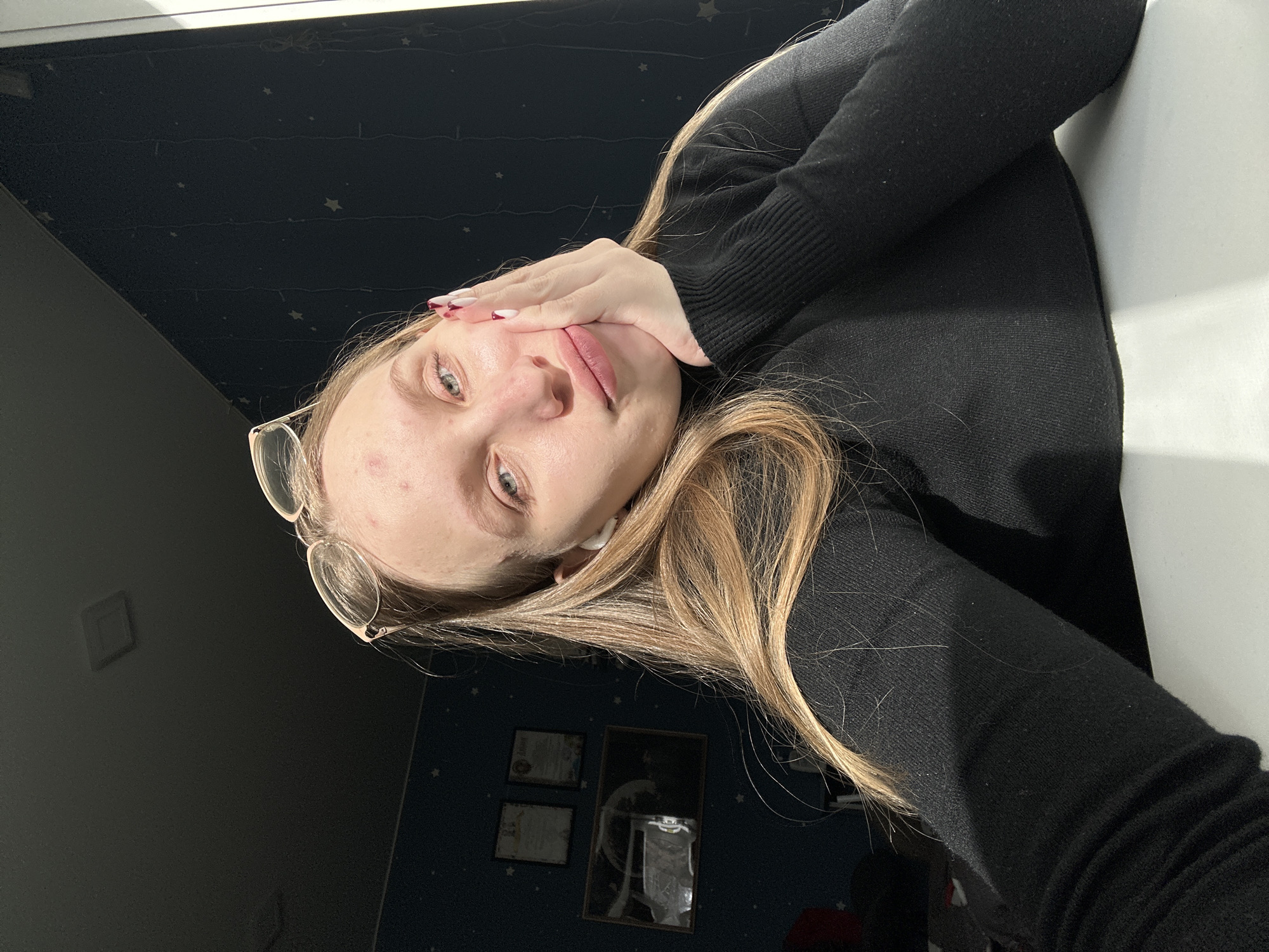 A young woman with long, light-brown hair and clear-framed glasses (worn on her forehead) is captured in a close-up, vertically oriented selfie. She wears a black long-sleeved top. Her left hand, adorned with white and red painted fingernails, gently touches her cheek, partially covering her mouth. She looks directly at the camera with a soft, pensive expression. Her skin shows some blemishes.
The background features a dark blue wall patterned with small white stars, creating a celestial theme. Two framed items, possibly certificates or small pictures, are visible on this wall, though their text is illegible. A bright, directional light source from the top-right illuminates the scene, casting soft shadows. To her right is a plain white surface, likely a wall or furniture. The setting is indoors, suggesting a personal space such as a bedroom or study. The overall impression is one of quiet contemplation in a distinctively decorated room in Chaikovsky, Russia.