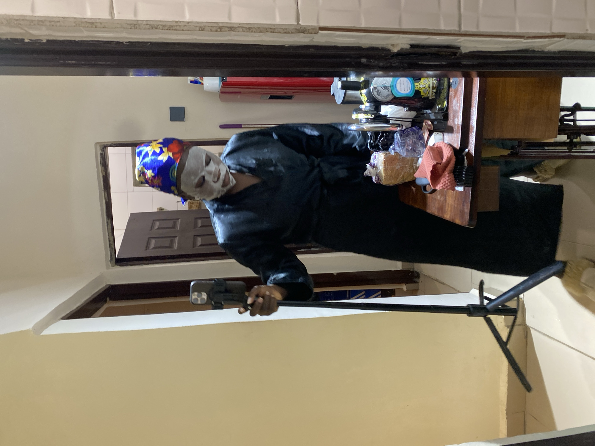 The image shows a person wearing a face mask and a blue floral headscarf in what appears to be an indoor environment. The person is holding a smartphone mounted on a tripod or selfie stick. Behind the person is an open doorway leading to another room with a dark-colored door. To the side, there is a cluttered counter or shelf with various items, including toiletries, packaged food, and other objects. The background reveals tiled walls and beige-painted walls. It's not possible to confirm this is Jos, Nigeria, from visual information alone. The main action is the person recording or taking a picture with their smartphone.