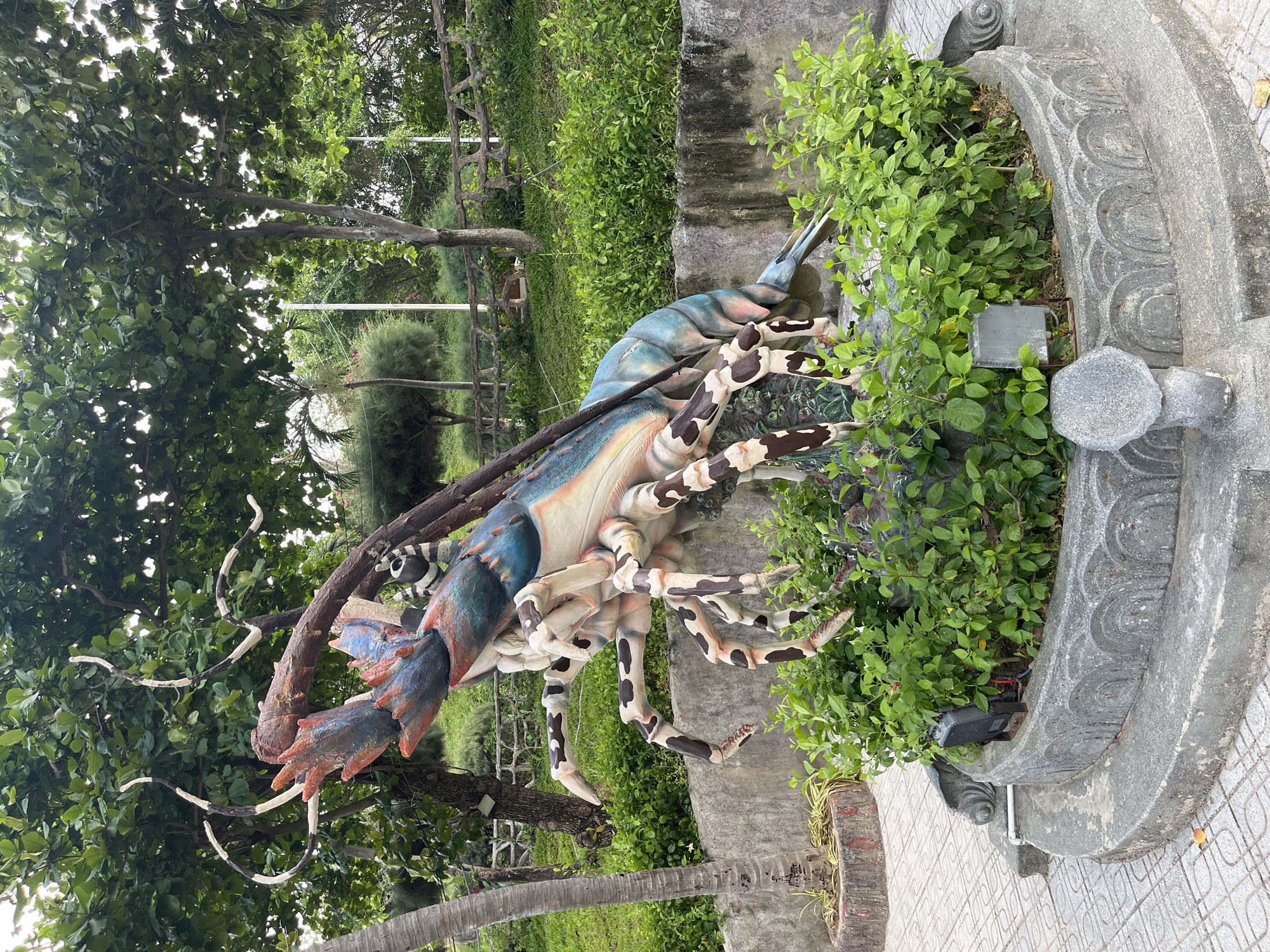 The image shows a large, colorful sculpture of a lobster in a park-like setting, likely in Bac Nha Trang Ward, Vietnam. The sculpture is positioned next to a concrete structure decorated with ornamental patterns and lush green plants. The sculpture's colors are a mix of blue, white, and shades of brown, with distinctive black spots along its legs. In the background are trees and glimpses of green grass and a fenced area. The sculpture is partially obscured by green foliage, creating a contrast with the surrounding vegetation. The ground is tiled with a geometric pattern, suggesting a well-maintained public space. The scene is well-lit, indicating outdoor daylight.