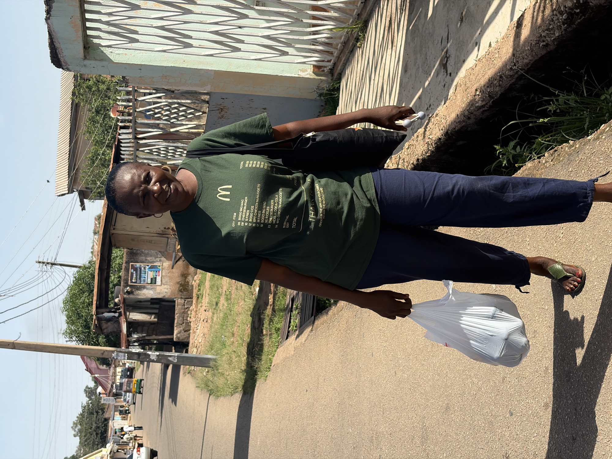 A woman stands on a city street holding a white plastic bag in one hand and several white objects in the other. She wears a dark green t-shirt with a McDonald's logo and a list of menu items and prices on the front, dark blue pants, and green sandals. A black bag hangs across her shoulder. The location appears to be in Jos, Nigeria, evidenced by the buildings and streetscape in the background. There is a fence behind her, and a concrete curb and storm drain to her right. In the distance, there is a street with other people, vehicles, and buildings.