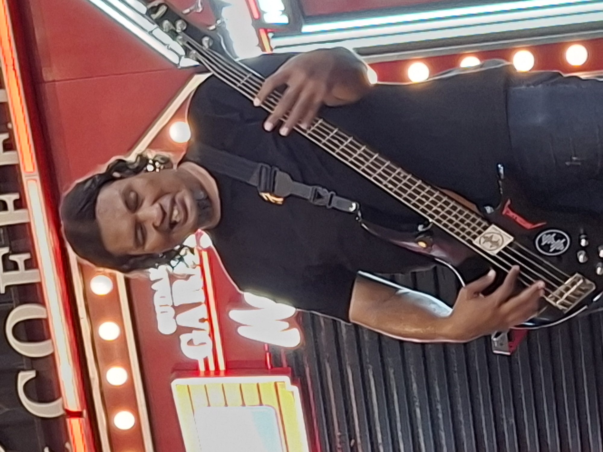 A medium shot captures an Indonesian man playing a black bass guitar in front of a red backdrop with neon signs. The man, with medium-length dark hair and a goatee, wears a black t-shirt and dark pants. His mouth is open as he plays, and his left hand is on the fretboard while his right hand strums the strings. The neon signs behind him are in Indonesian, with "COFFEE" visible at the top, and below it "GUDANG GARAM" and "MOJO". The background also features several illuminated lightbulbs. The scene appears to be outdoors, possibly at a venue or event in the city of Lhokseumawe, Indonesia.