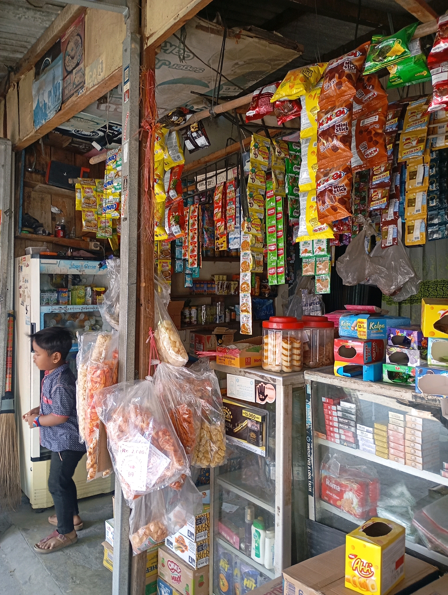 In Babelan, Indonesia, a young boy stands by the entrance of a small, open-air convenience store. He is dressed in a dark blue and white patterned shirt and black pants, with light brown sandals on his feet. The store is densely packed with merchandise, including shelves filled with snacks, drinks, and household items. Various colorful snack packets are hanging from the ceiling, and large clear plastic bags filled with snacks are displayed near the entrance. A refrigerator is visible behind the boy, containing more products. The store appears to be made of wood and corrugated metal, with a rustic and cluttered appearance, typical of many small businesses in the region.