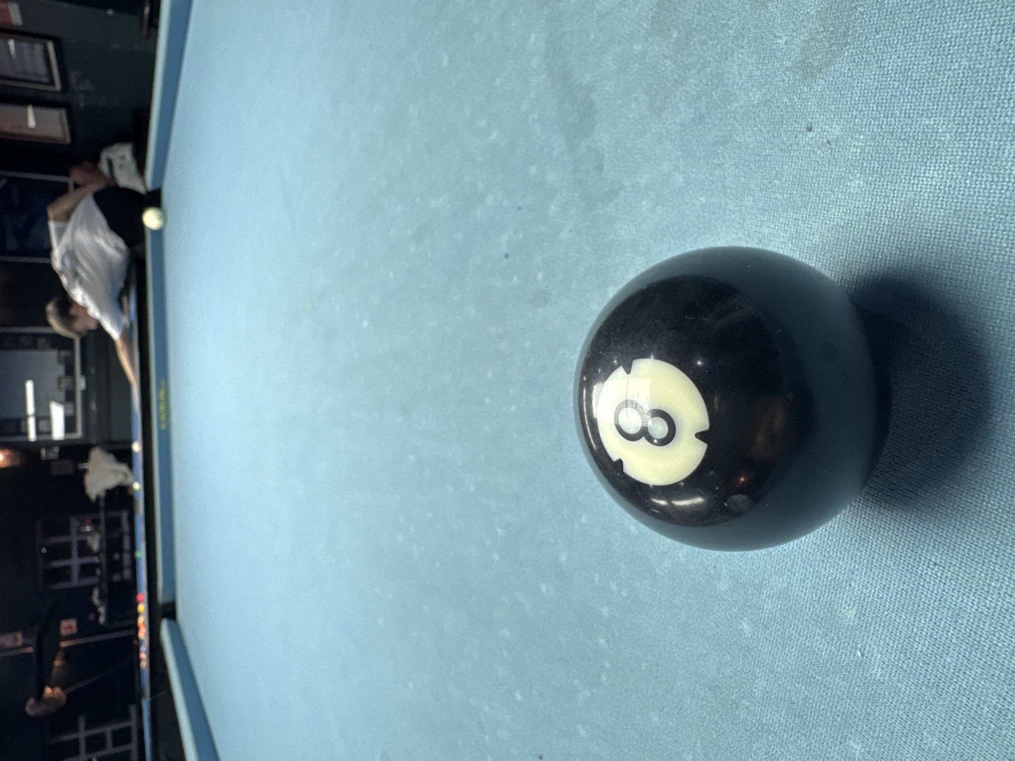 A billiards table, covered in light blue felt, is the primary focus. An eight ball sits prominently on the felt, casting a distinct shadow. A blurred figure of a person holding a pool cue is visible near the edge of the table. The background shows a portion of a bar or entertainment establishment, including tables, chairs, and a wall with framed pictures. There is no information to determine that it is located in Bellville, South Africa.