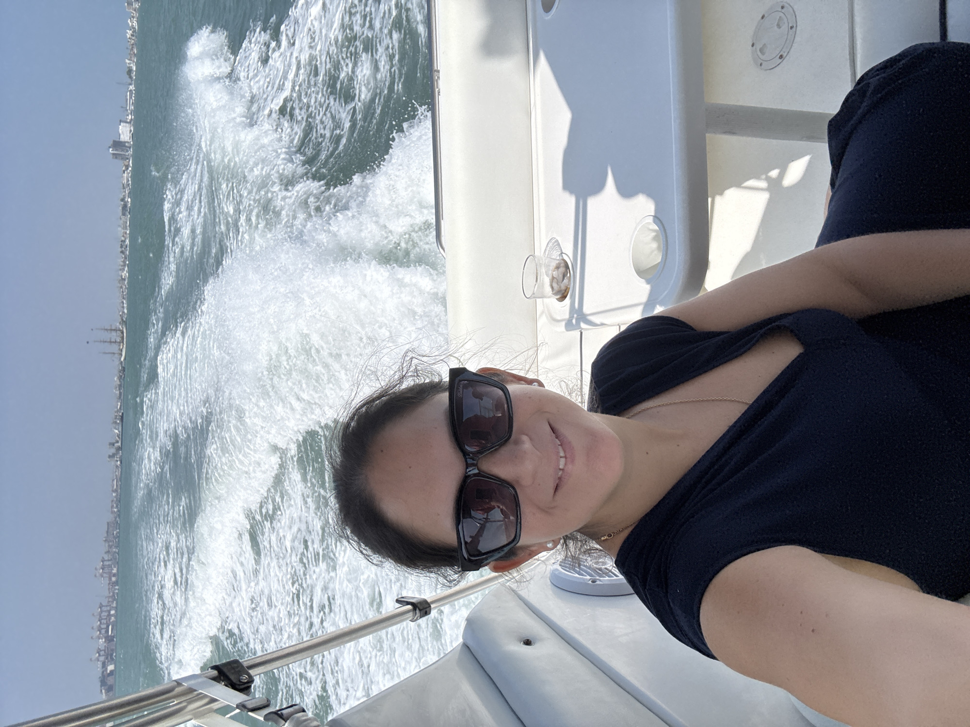 A woman wearing sunglasses and a black top is taking a selfie on a boat. The boat is moving, as evidenced by the large wake trailing behind it. In the background, a cityscape is visible. Given the prompt, this is likely La Punta, Peru. A clear plastic cup with its content is visible next to a white table.